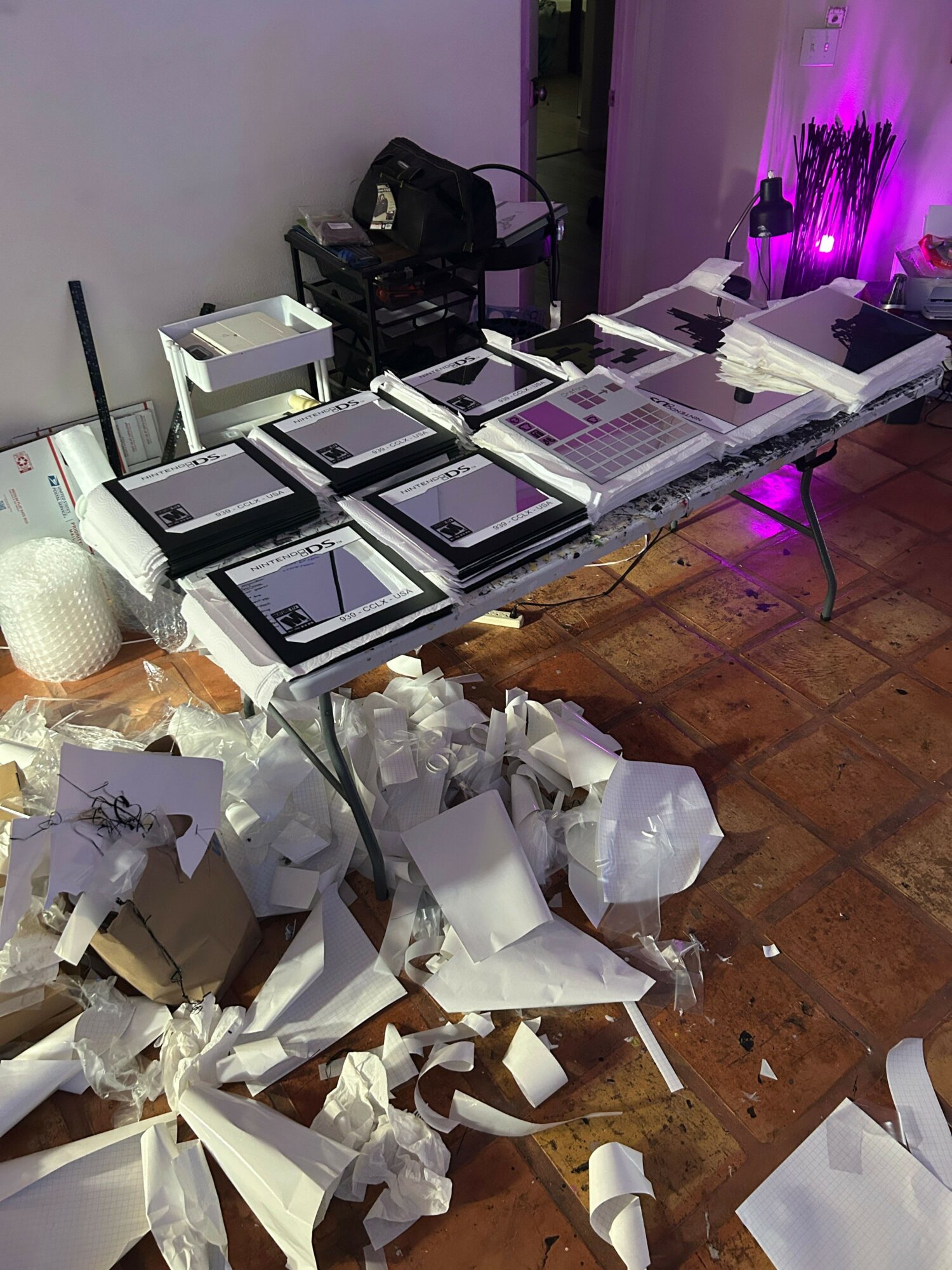 Table with electronic devices and papers, cluttered with torn paper and packaging on the floor, purple lighting in background.