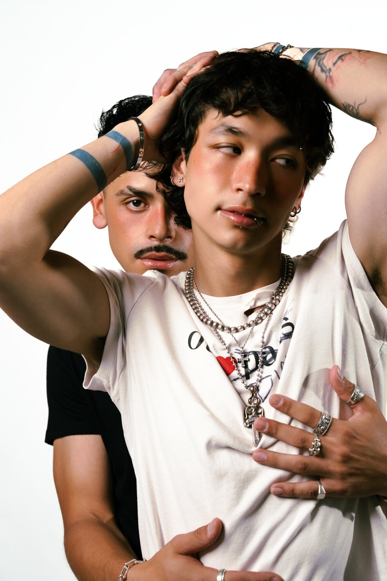 Two young men pose closely, one with hands on head and the other with hand on chest, both wearing jewelry.