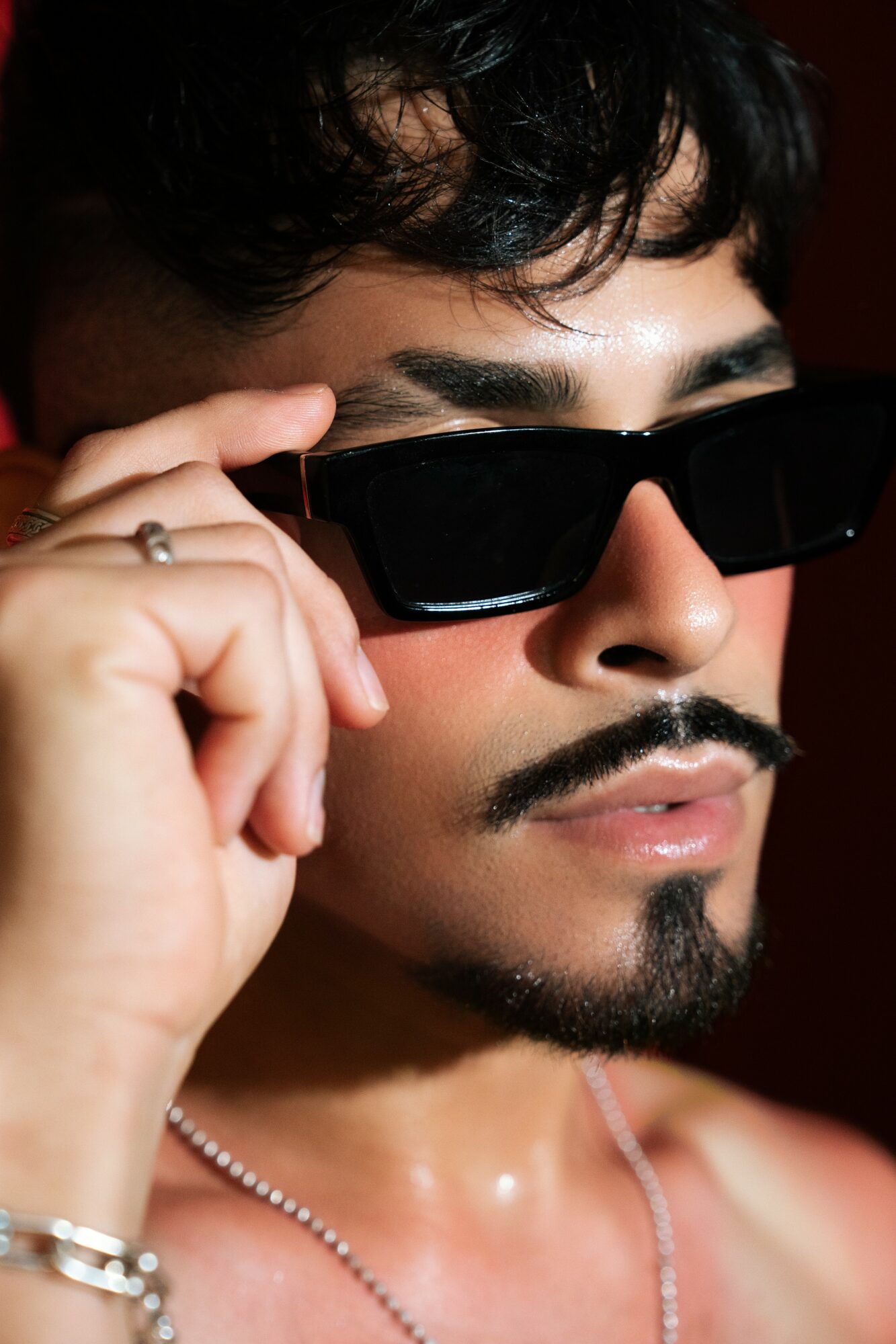 Close-up of a person with dark hair, sunglasses, and a mustache, adjusting their glasses with a hand, against a dark background.
