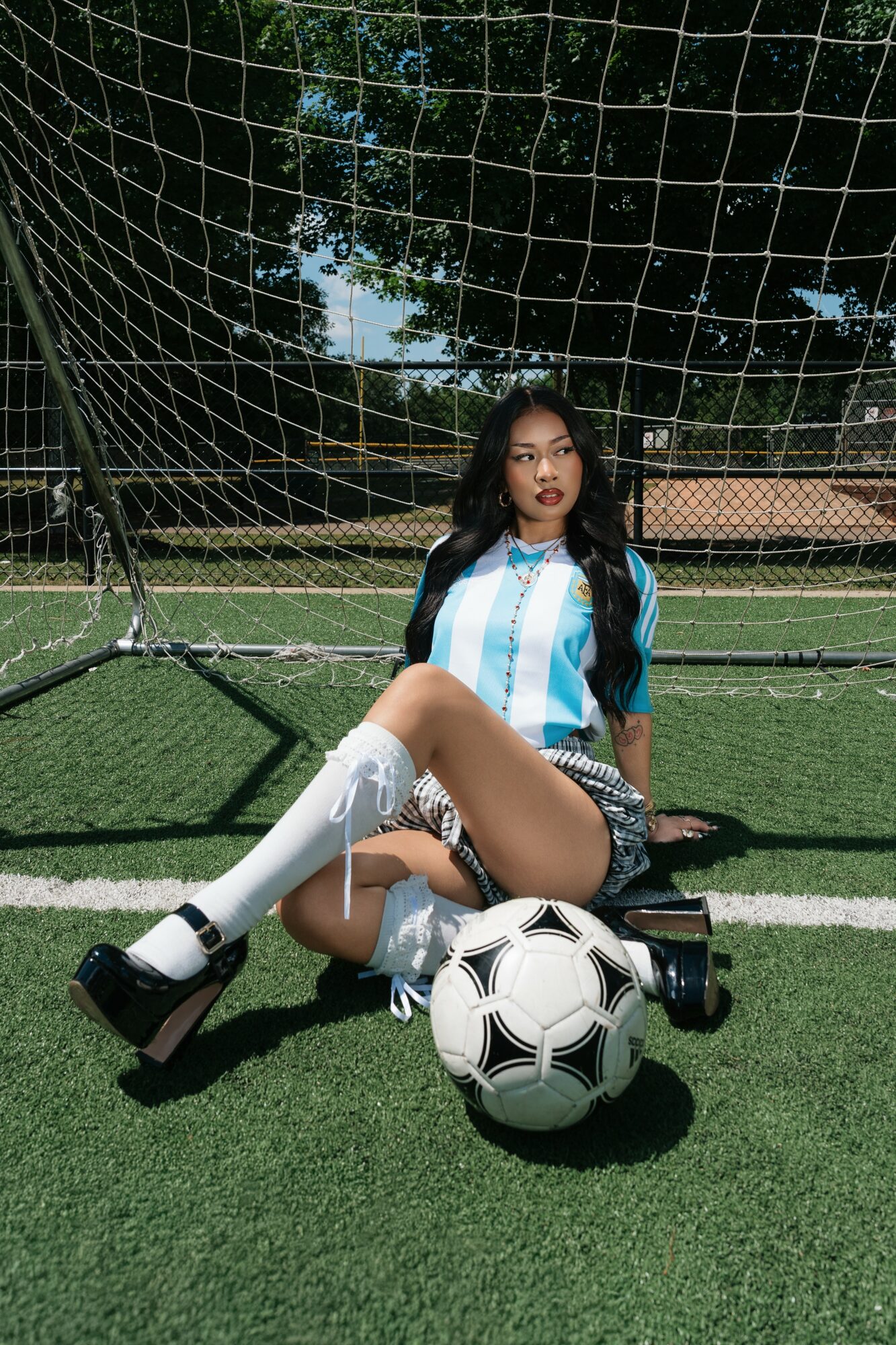 Person sitting on artificial grass soccer field with soccer ball, wearing striped shirt, shorts, and knee-high socks, against a goal net background.