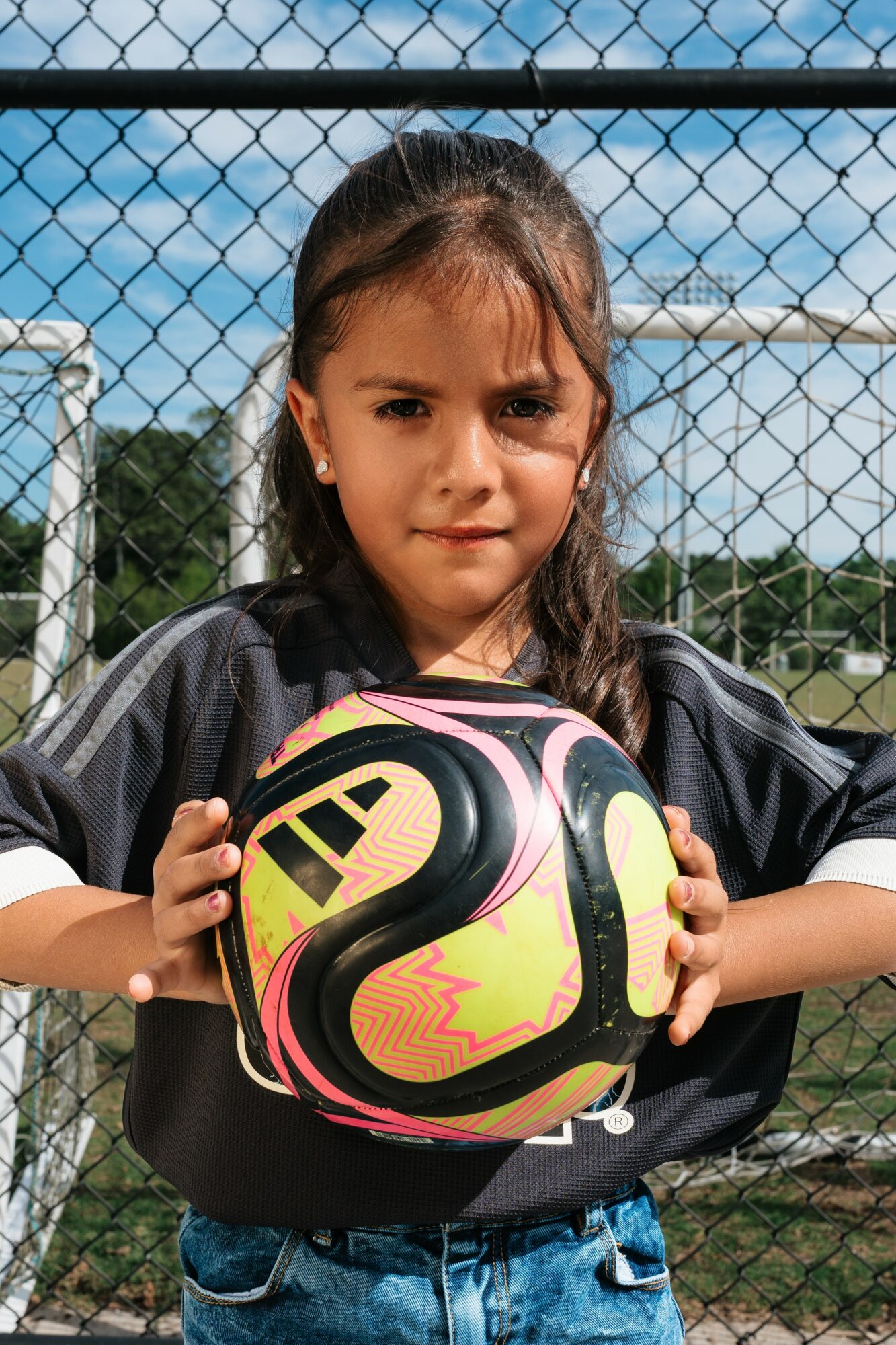 Young girl holding a colorful soccer ball outdoors, standing in front of a chain-link fence with a blue sky background.
