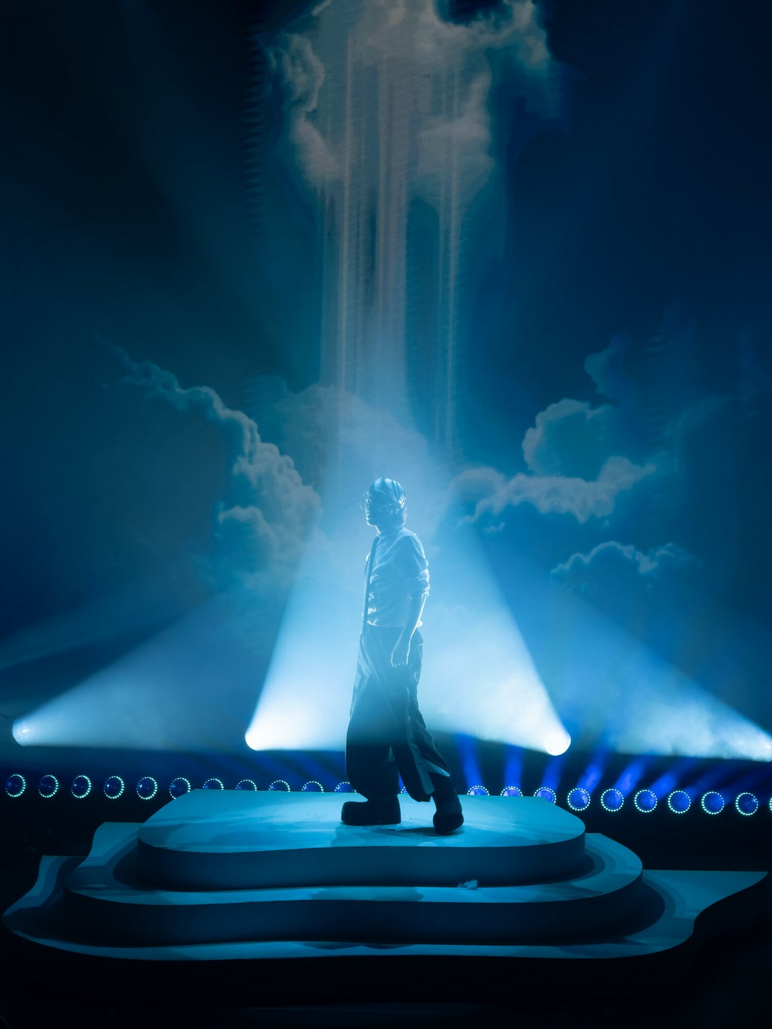 Person standing on a stage with dramatic lighting and clouds in the background.