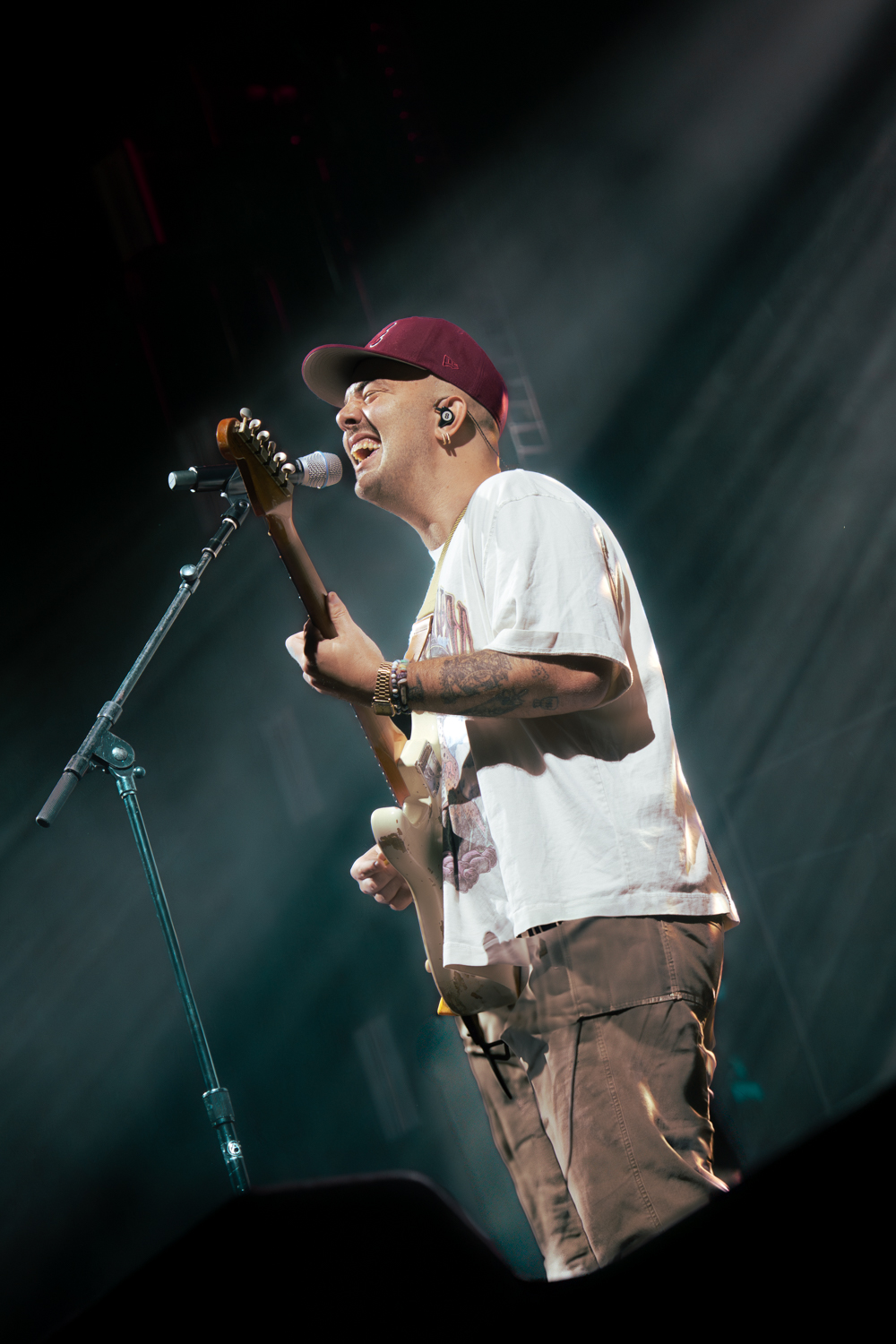 Person wearing a maroon cap and white t-shirt playing an electric guitar on stage with spotlight beams.