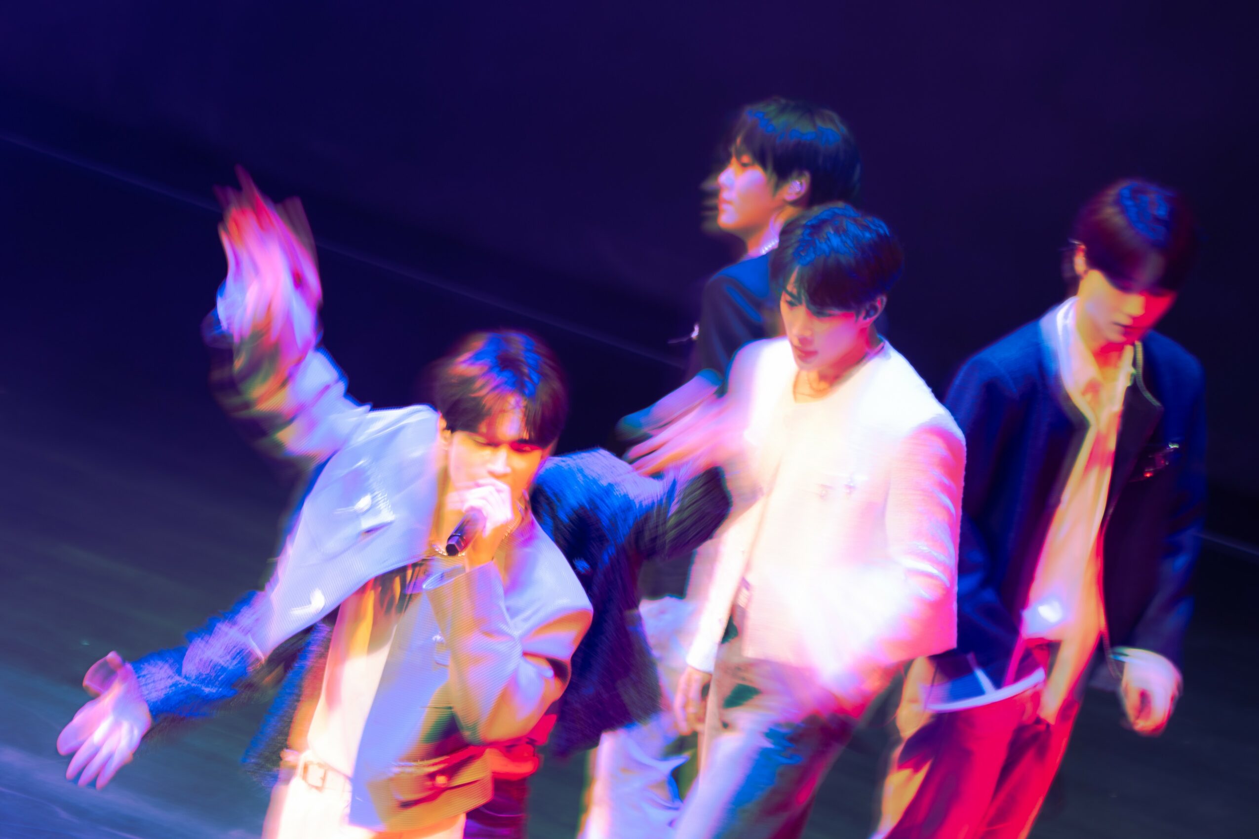 Five young men perform on stage with dark background, wearing stylish clothing, some holding microphones, in a dynamic pose.