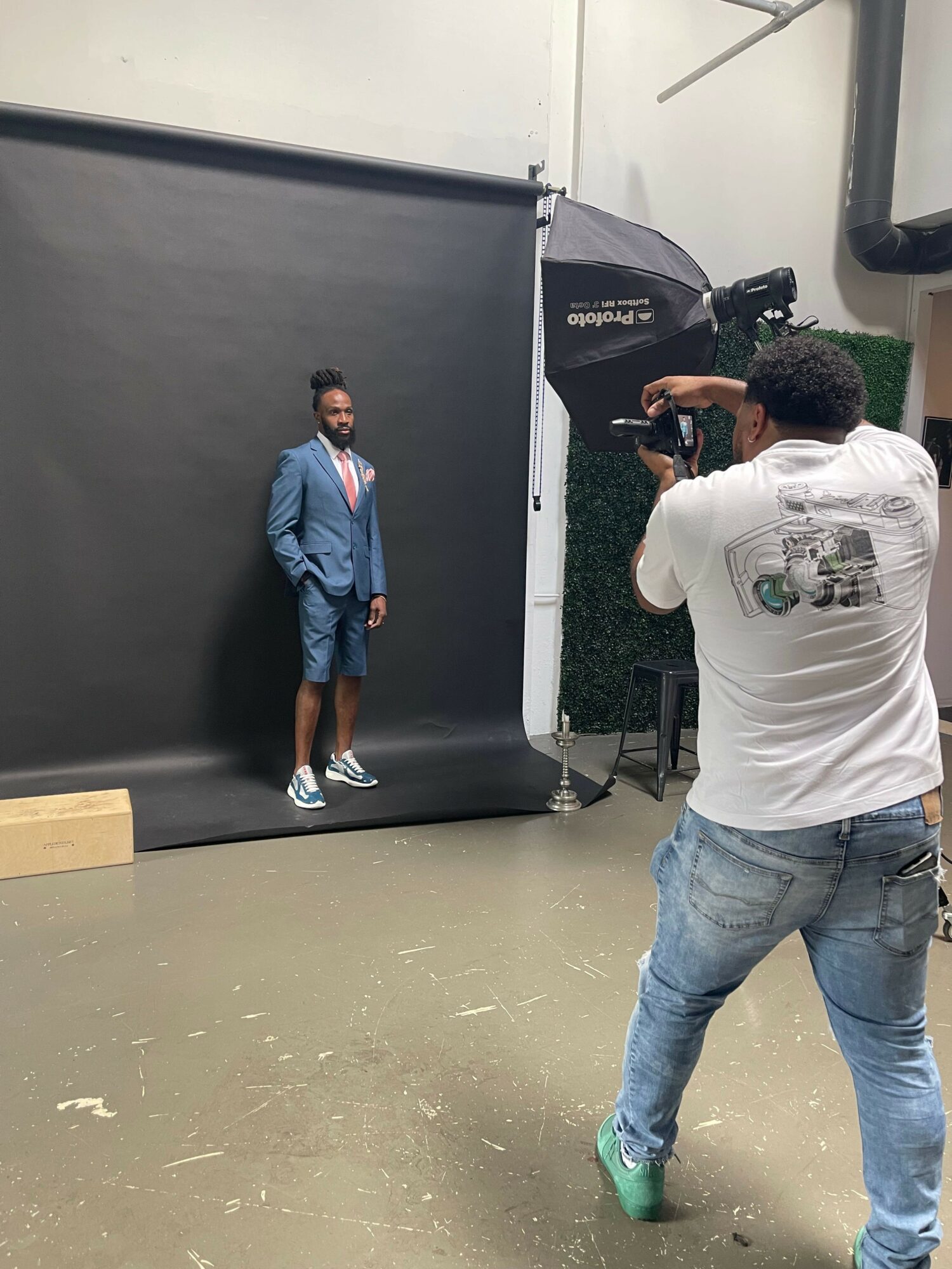 Person in white shirt taking a photo of a person in blue jacket standing in front of a black backdrop.