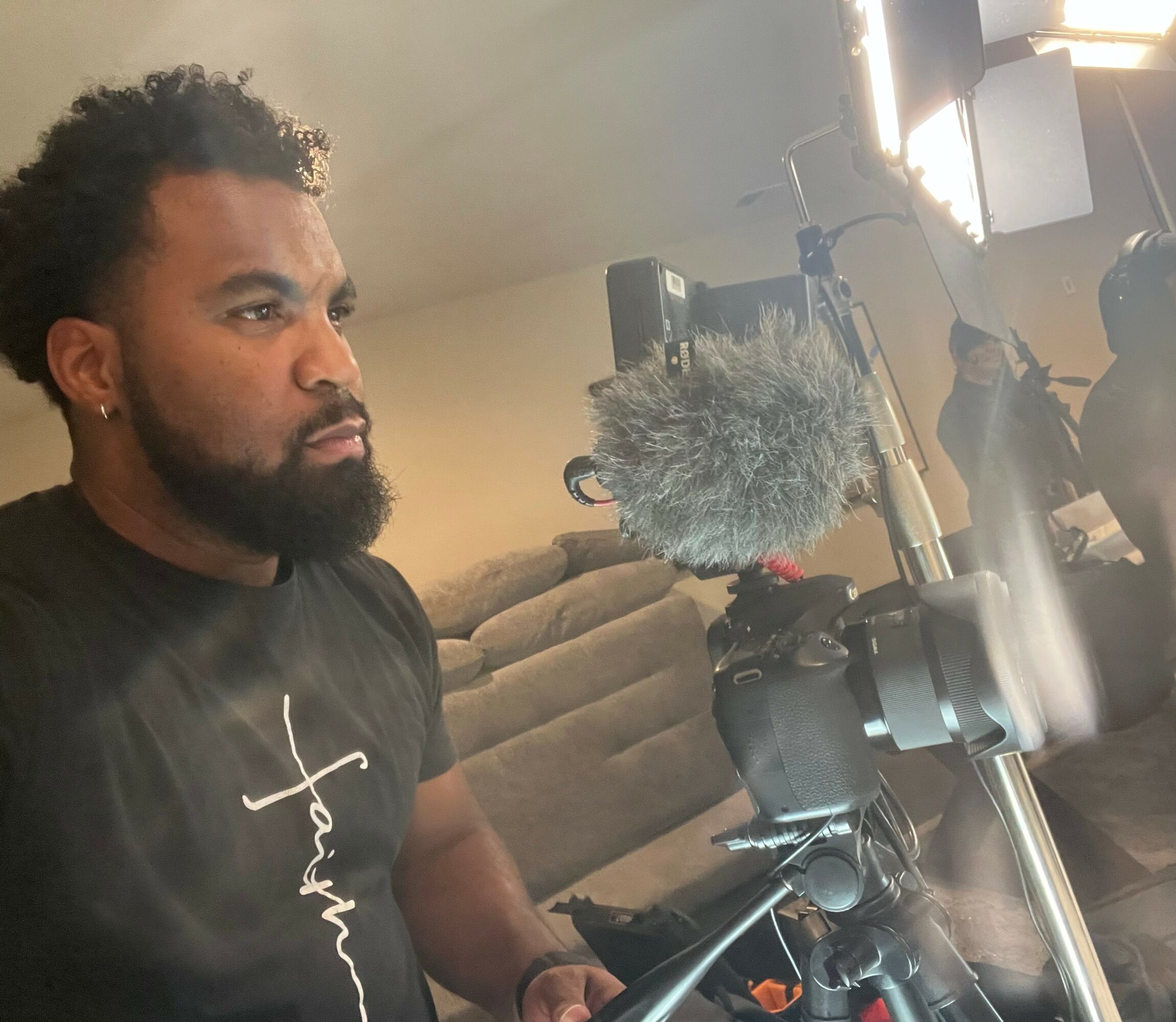 Man with curly hair and beard operating camera with microphone, in indoor setting with sofa and lighting equipment.