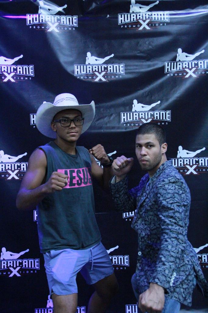 Two young men pose in front of a black backdrop with 'HURRICANE' logos, one wearing a hat and glasses, both with fists raised.