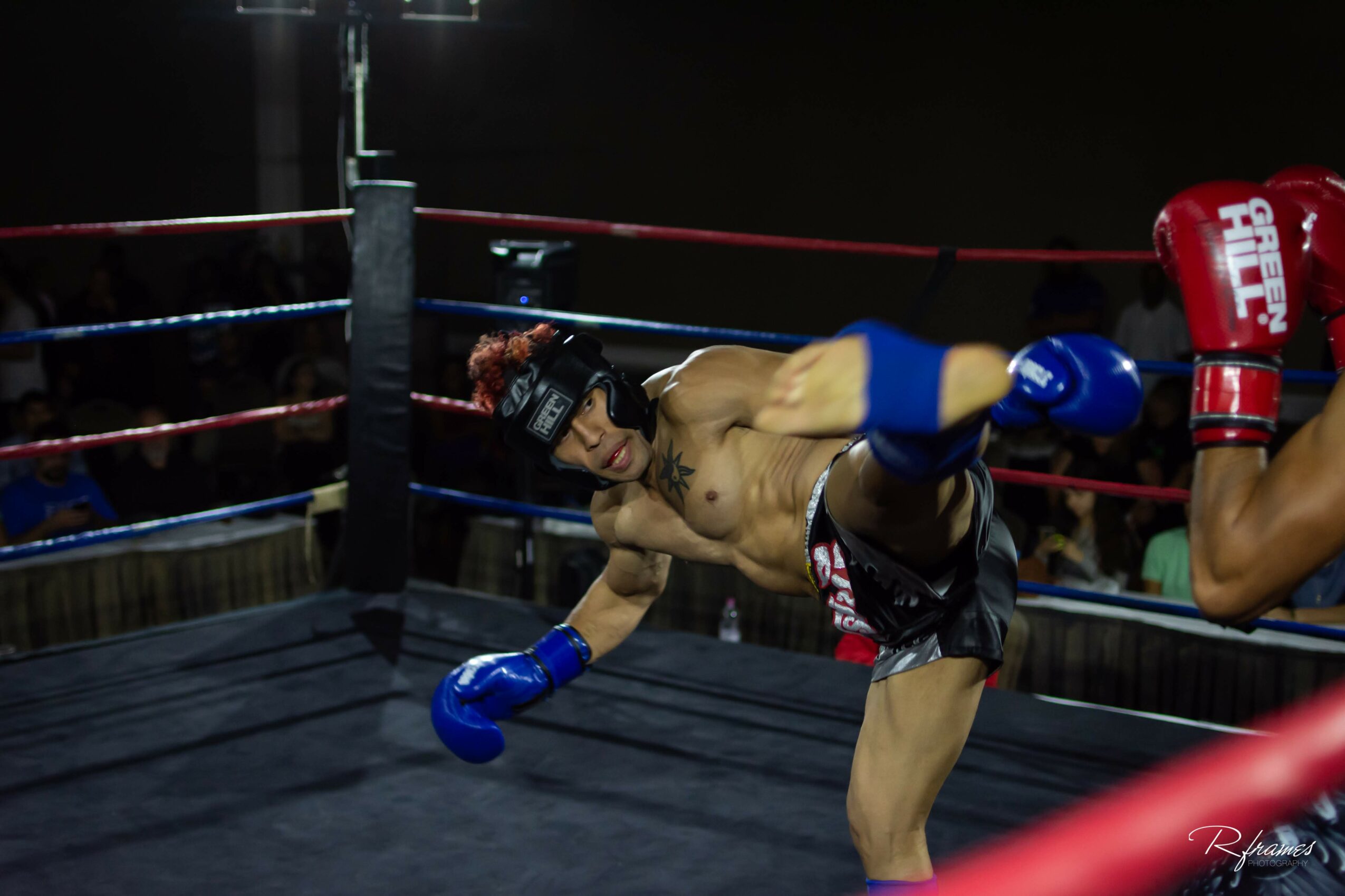 Muay Thai fighter in blue gloves and shorts kicks an opponent in a boxing ring, with ropes and audience visible.