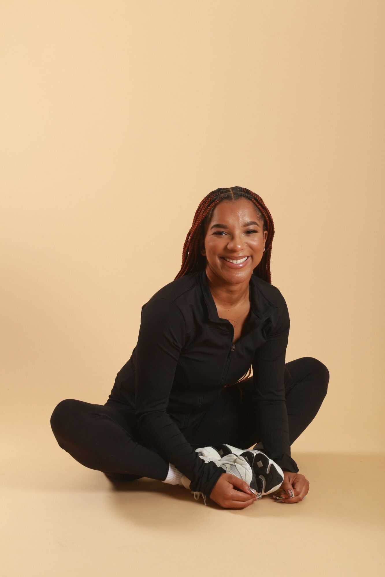 Person sitting on the floor against a plain wall, smiling, with legs crossed and hands resting on knees.