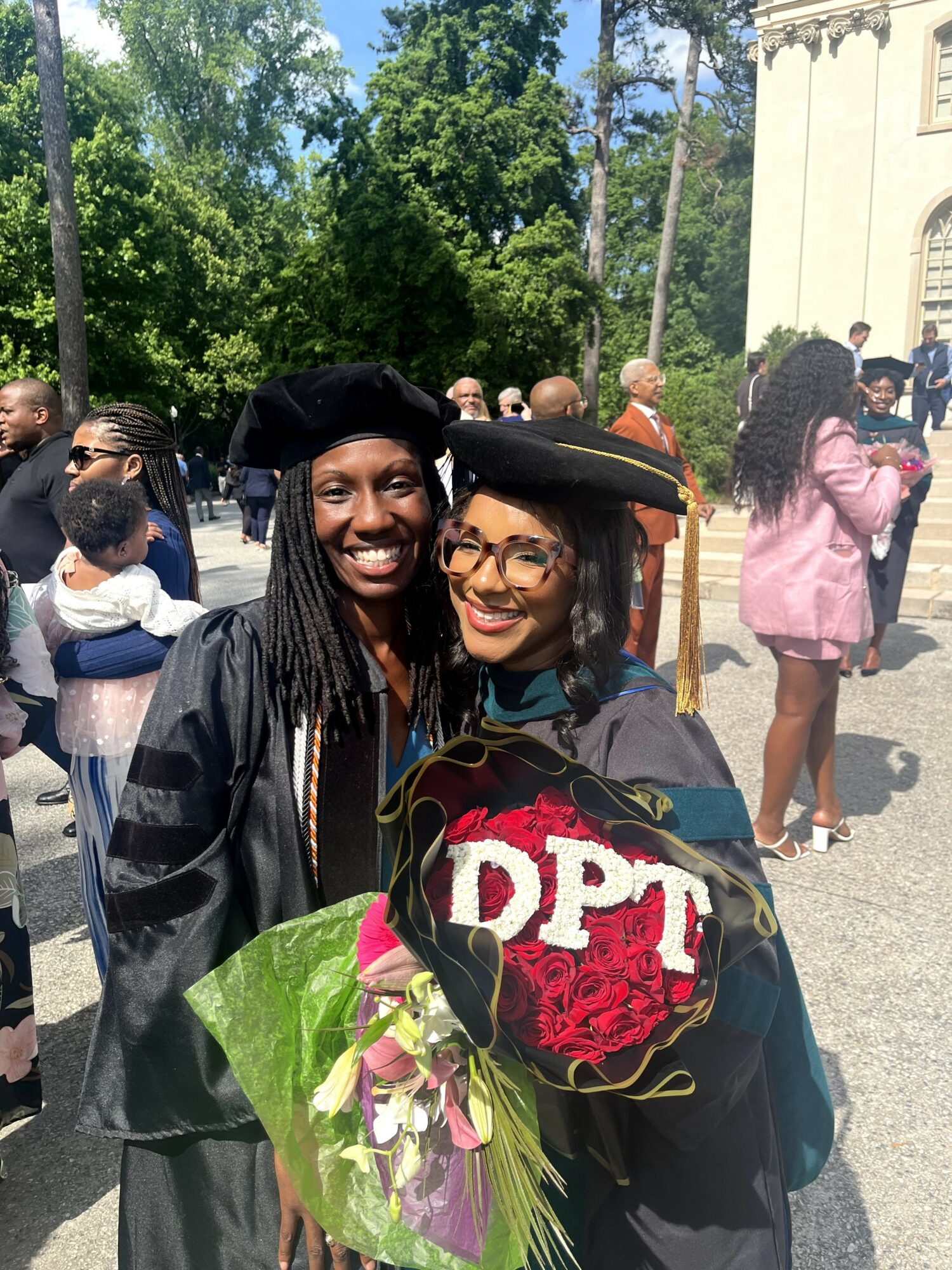 Two women in graduation caps and gowns smiling, holding a bouquet of flowers, outdoors with trees and people in background.