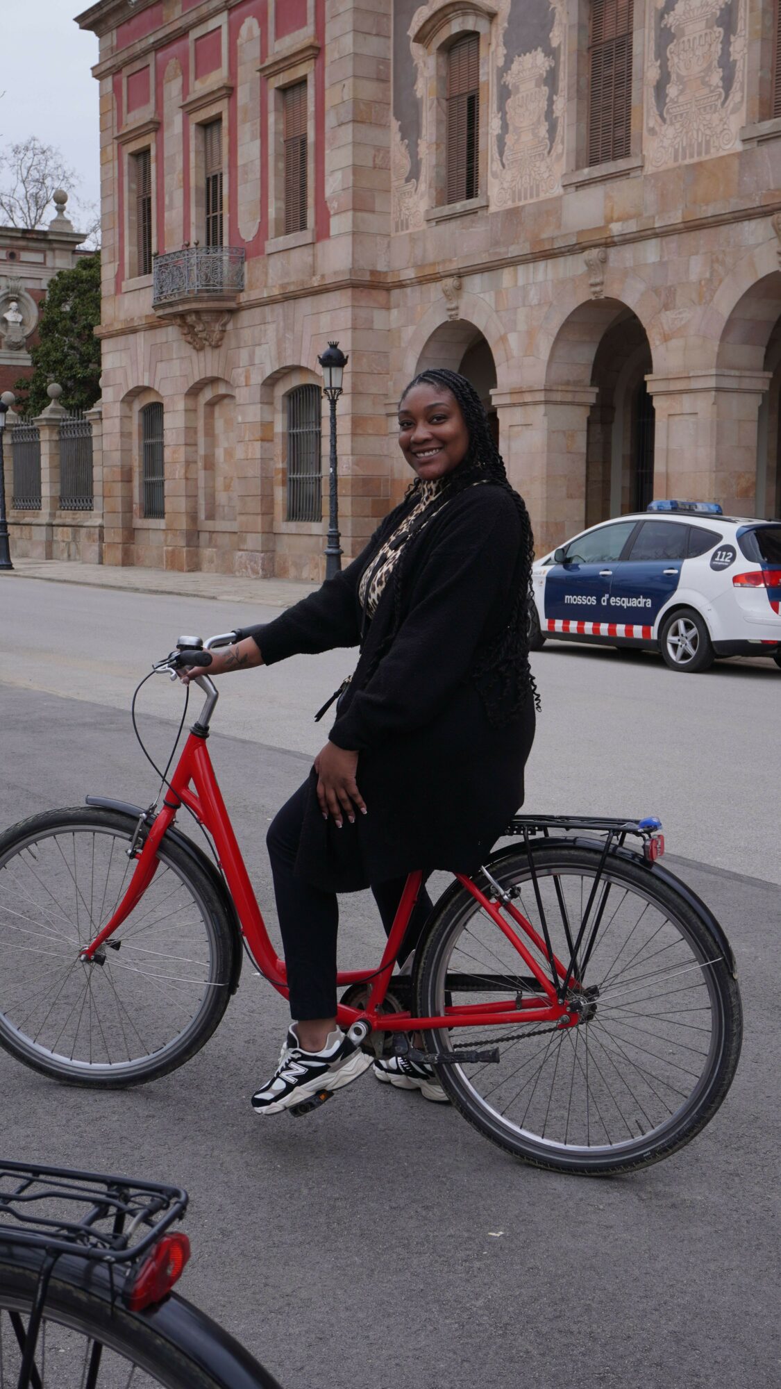 Person standing next to a red bicycle on a city street, holding a phone, with buildings and a car in the background.