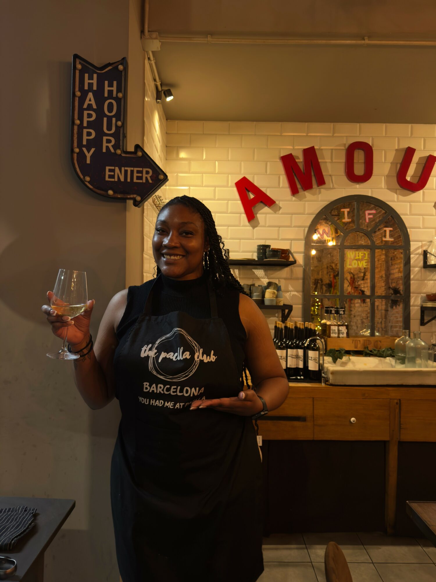 Woman holding a glass of wine in a restaurant with red letters spelling 'AMOUR' on the wall behind her.