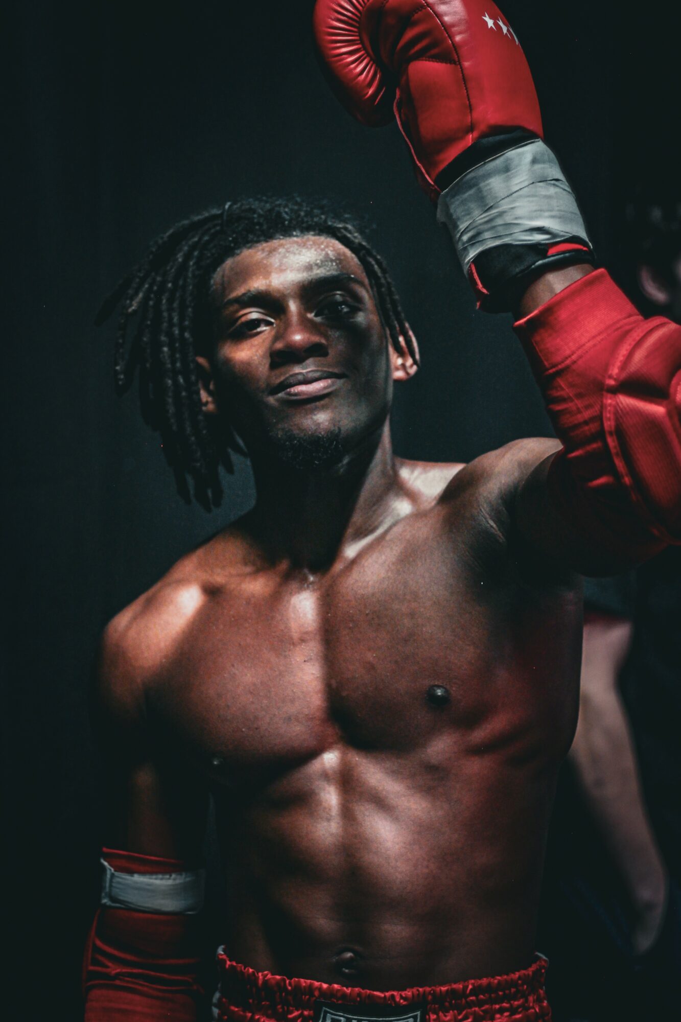 Boxer with gloves raised, shirtless, muscular, dark background, confident expression, dreadlocks, in a boxing stance.