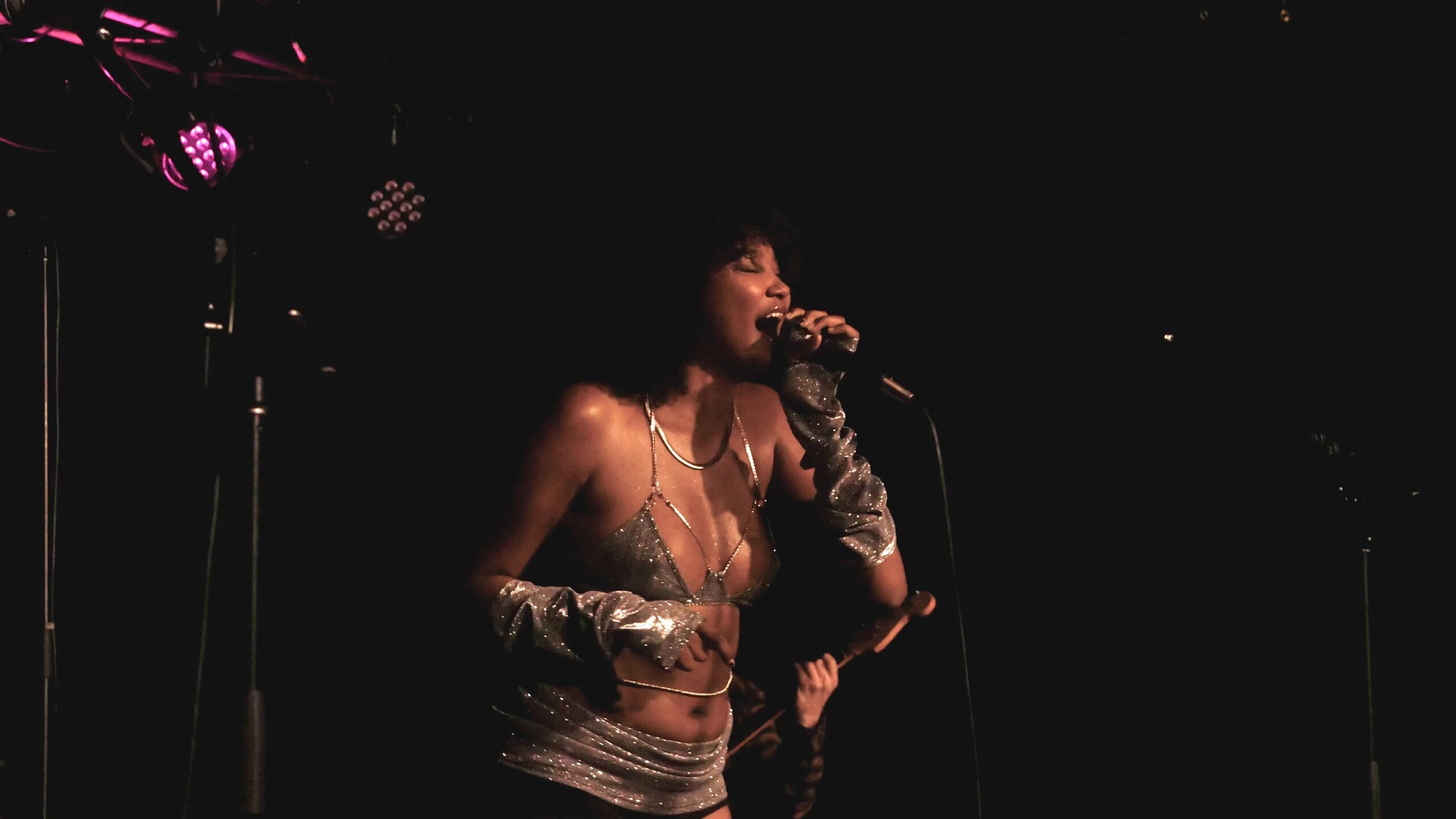 Person singing into a microphone on stage with dark background, wearing a shiny top and gloves.