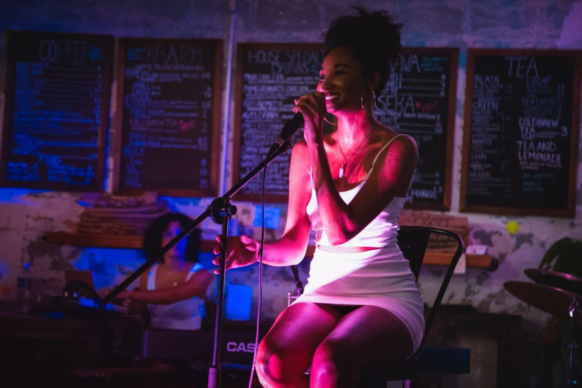 A woman singing into a microphone on stage with a dark background and colorful lighting, wearing a white dress.