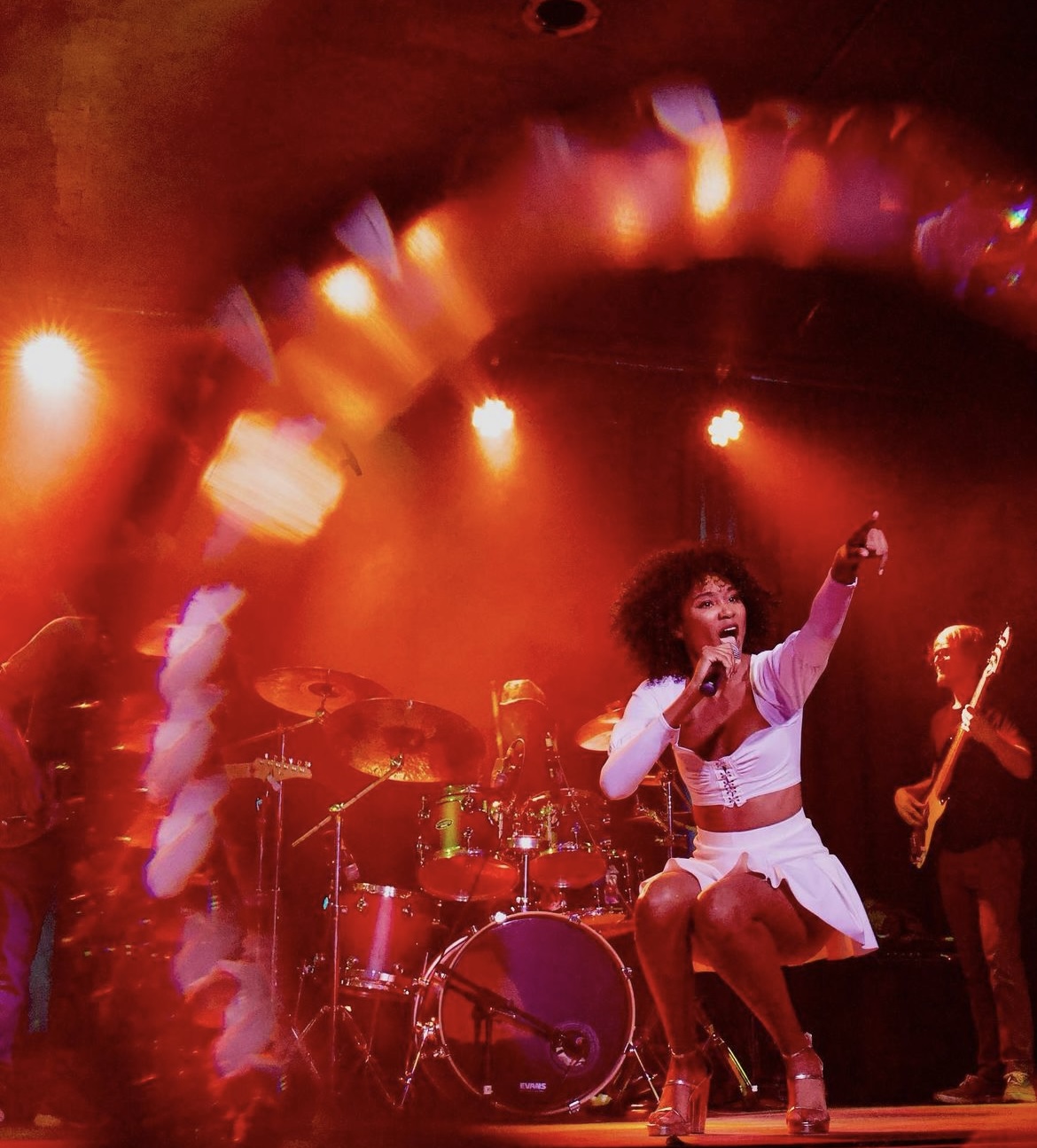 Performer singing on stage with red lighting, drums, and a guitarist in the background.