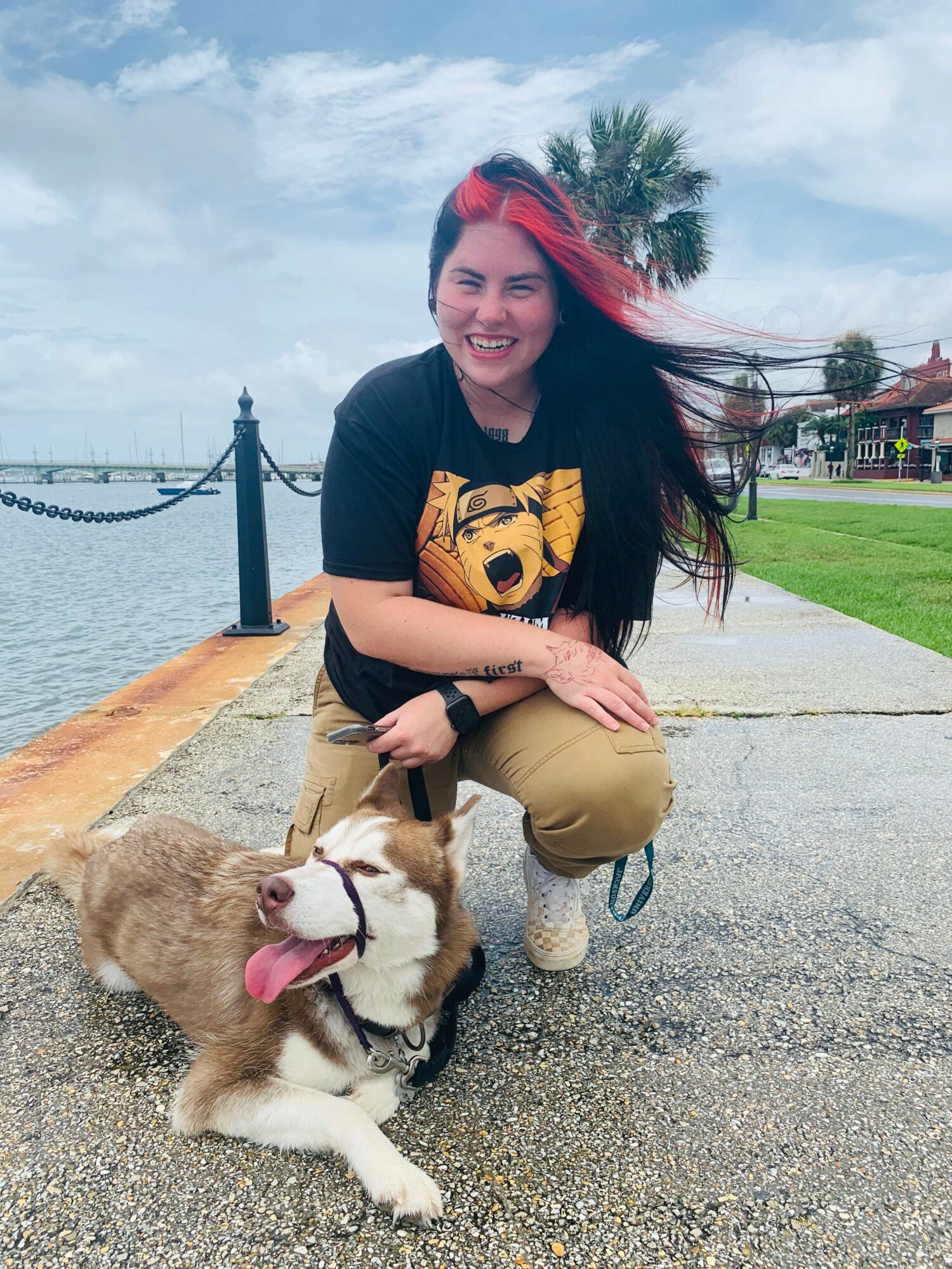 Woman crouching outdoors with a smiling dog near water, palm trees, and a cloudy sky in the background.