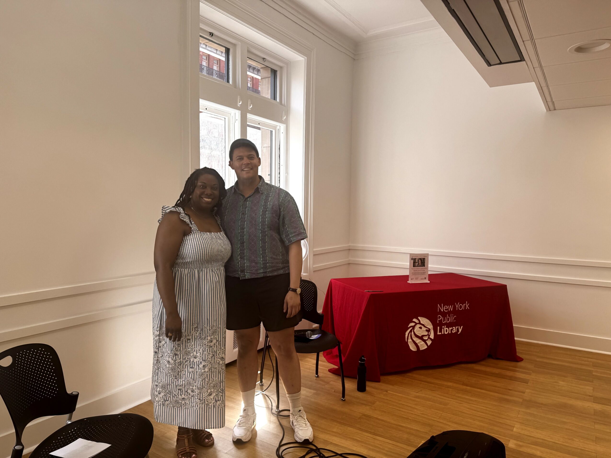 Two people standing together in a room with a window, a table with a red cloth, and chairs.