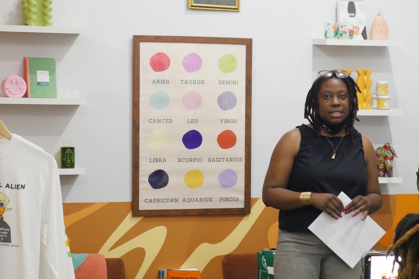 Woman standing in front of a colorful chart on a white wall, holding papers, with shelves and decorations around.