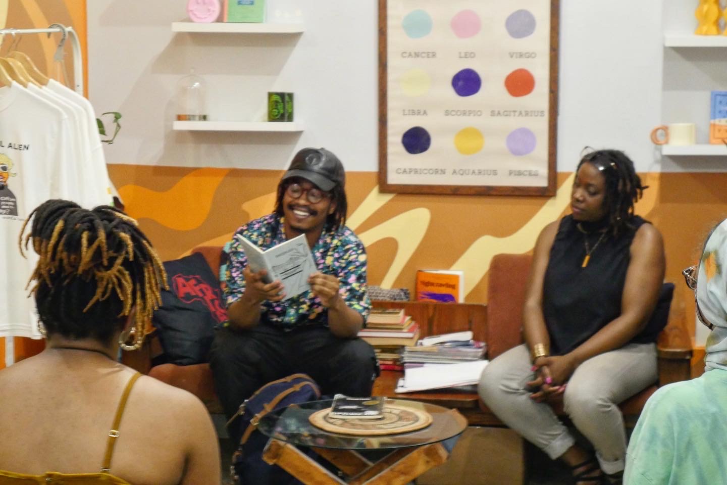 Group of women sitting in a room, one woman speaking and holding a book, others listening, shelves with books and posters behind them.