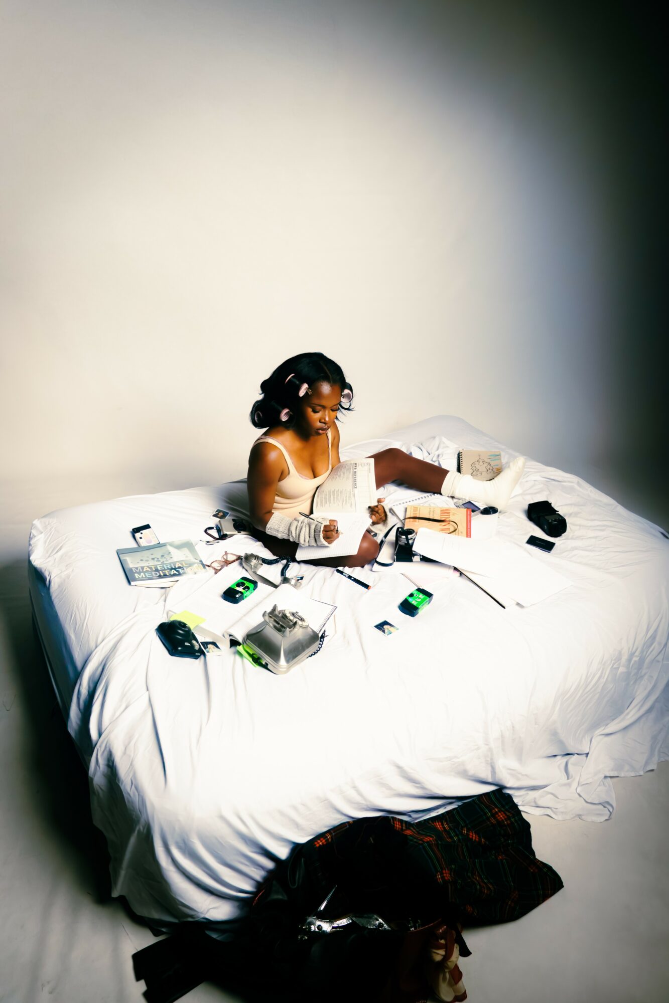 Person sitting on a bed surrounded by electronic devices and papers, with a plain wall background.