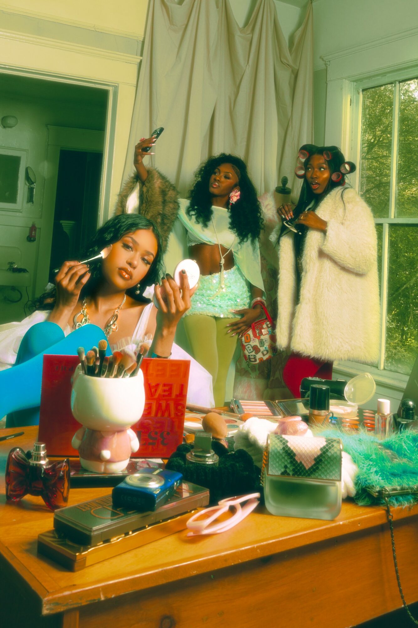 Four women in a room with makeup and beauty products on a table, one applying makeup, others standing near a window.