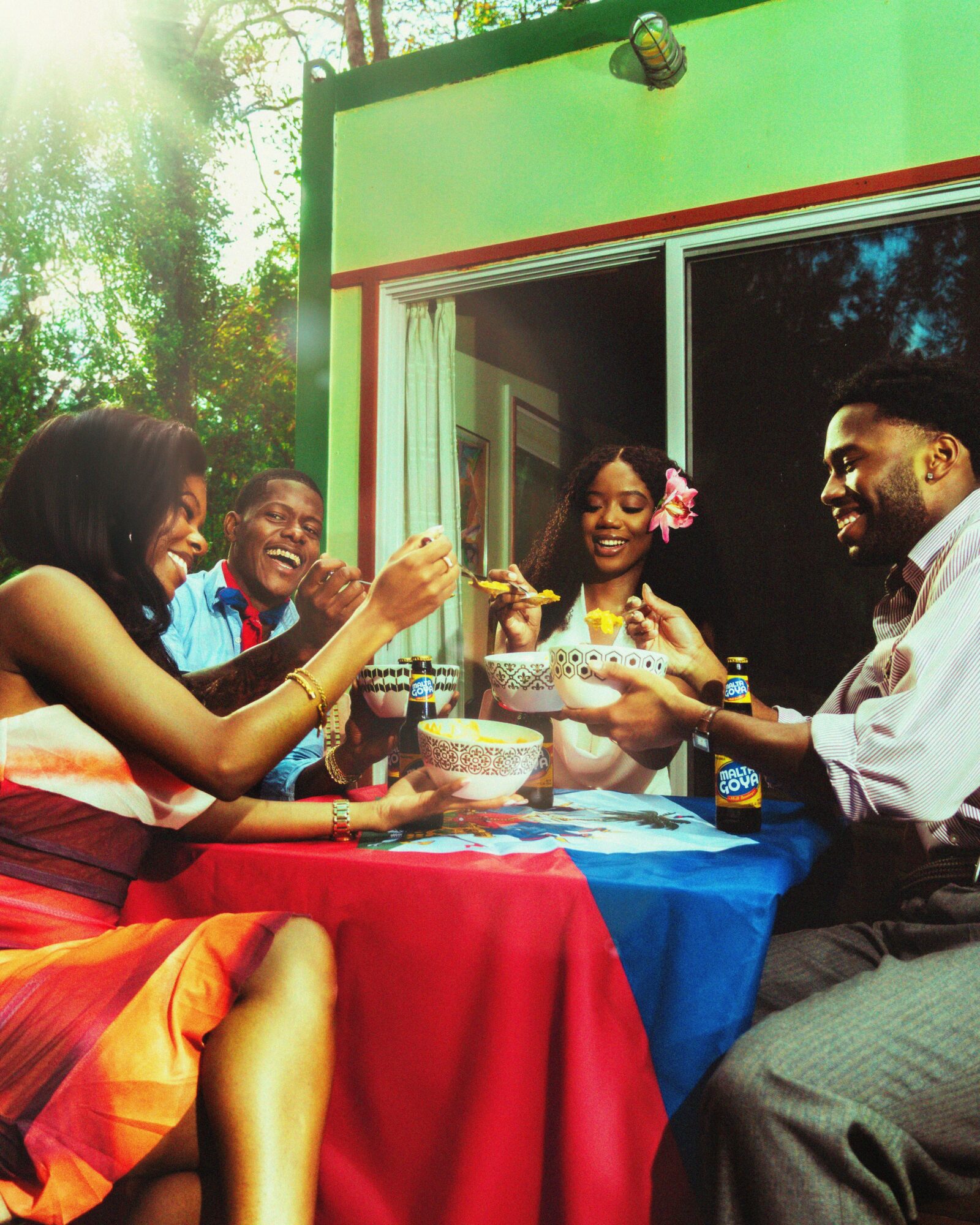 Four people sit around a table outdoors, enjoying food and smiling, with a green building and trees in the background.