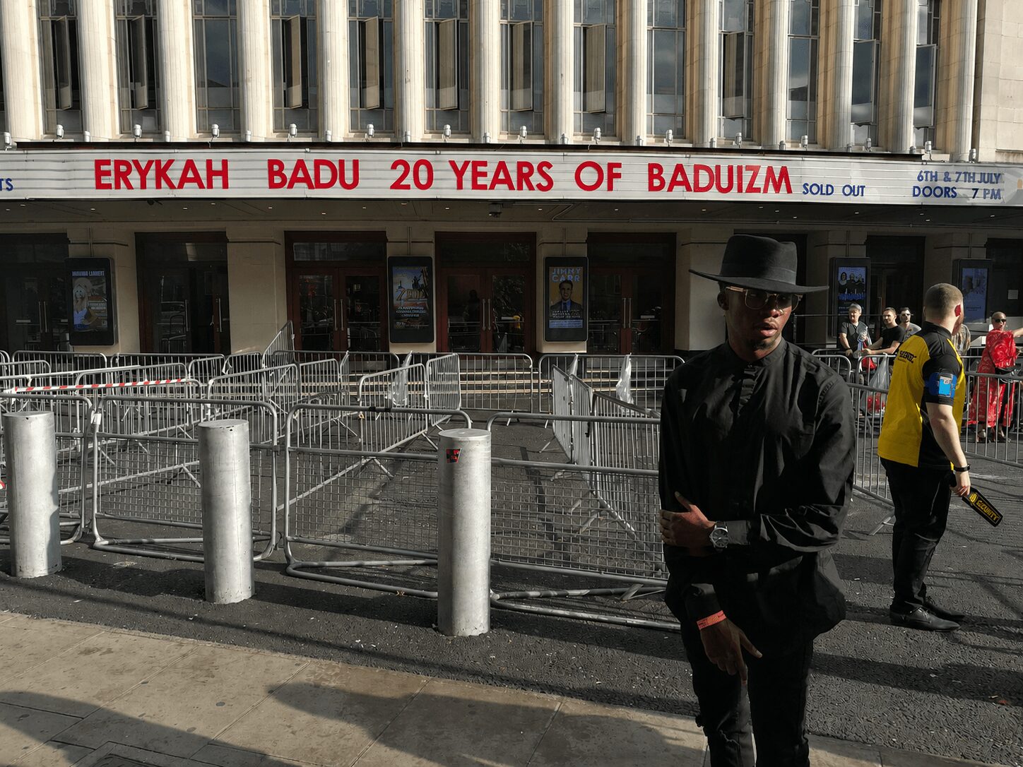 Person wearing black clothing and a hat standing outside a building with a sign about 20 years of Baduizm, surrounded by metal barriers.
