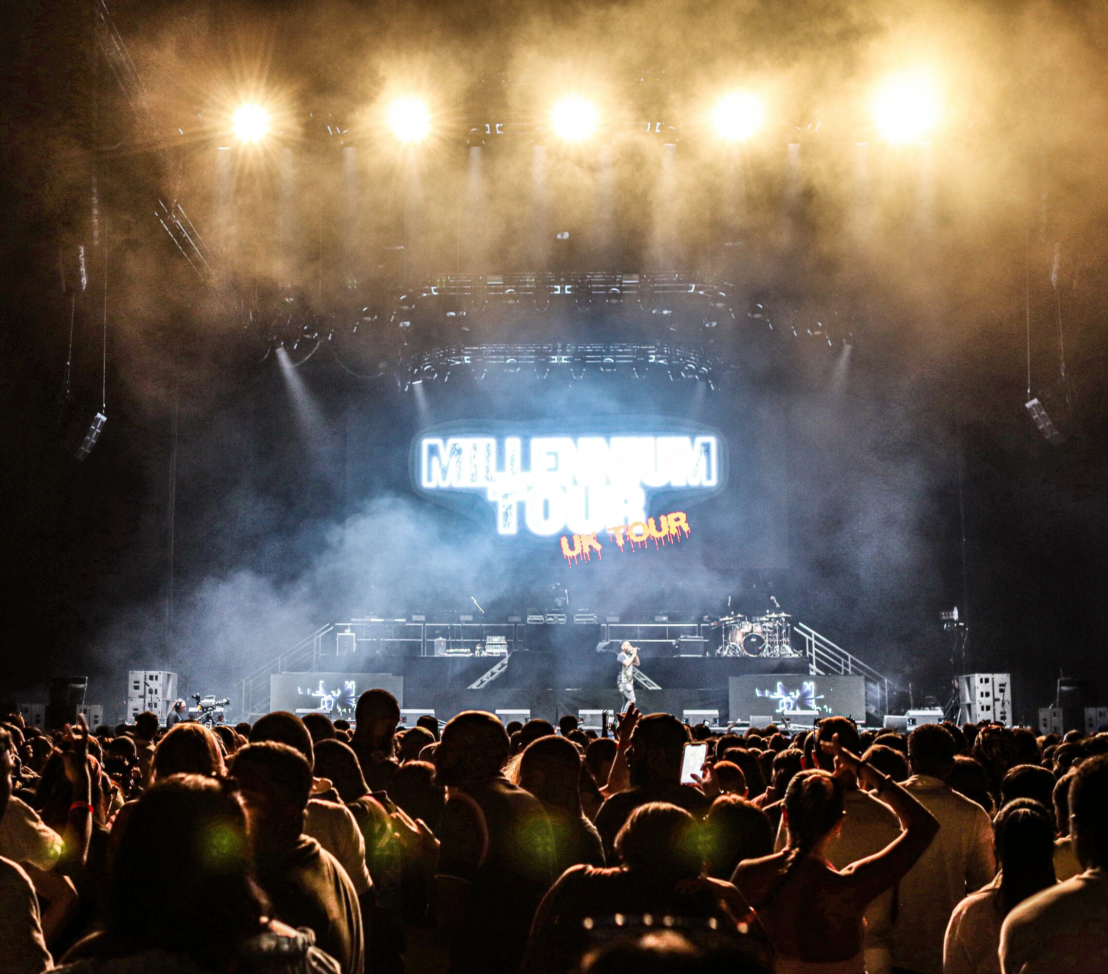 Concert stage with bright lights, fog, and a large screen, audience watching the performance.