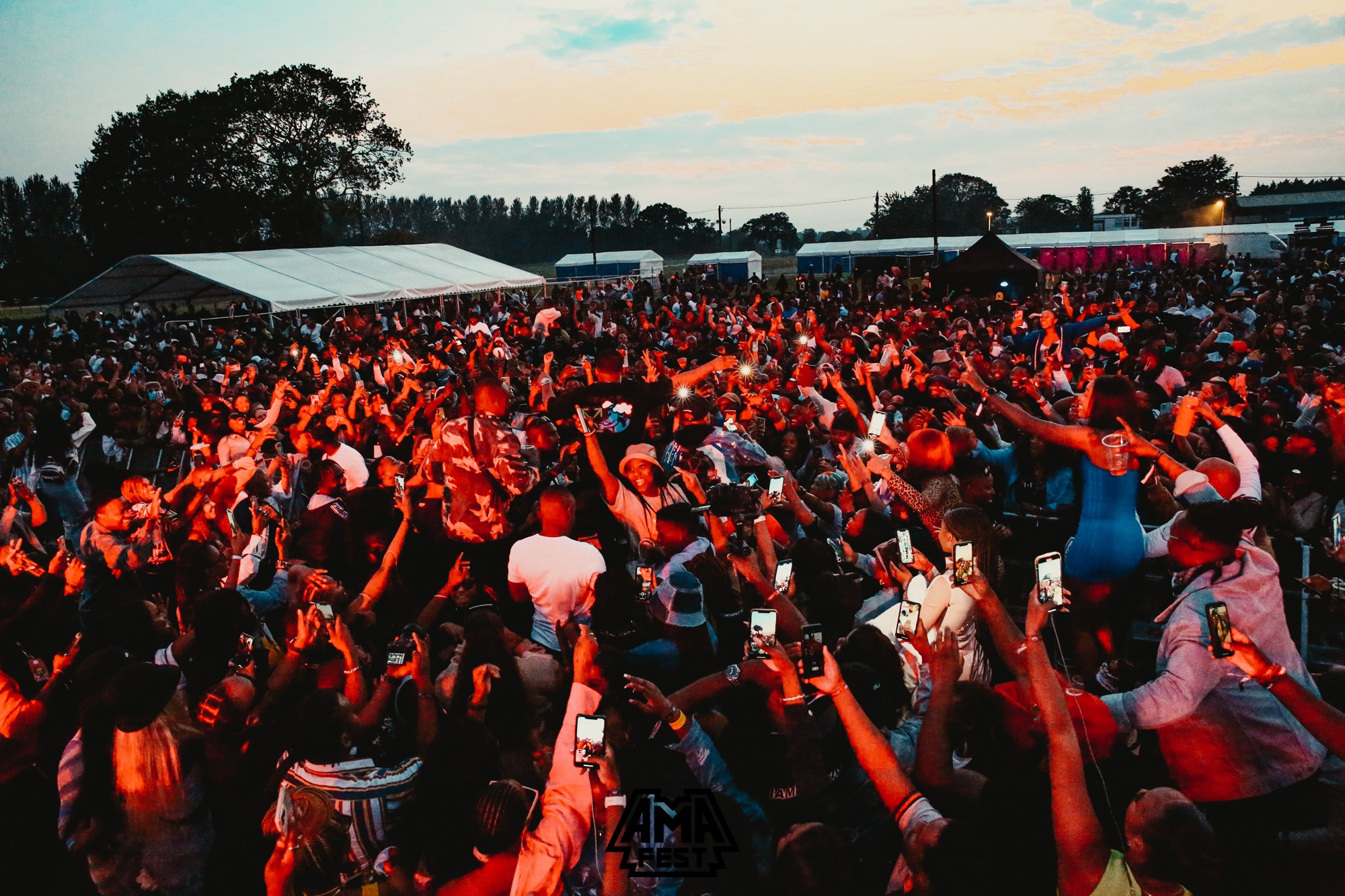 Large crowd at an outdoor event during sunset, with many people raising their hands, some holding phones, and tents in the background.