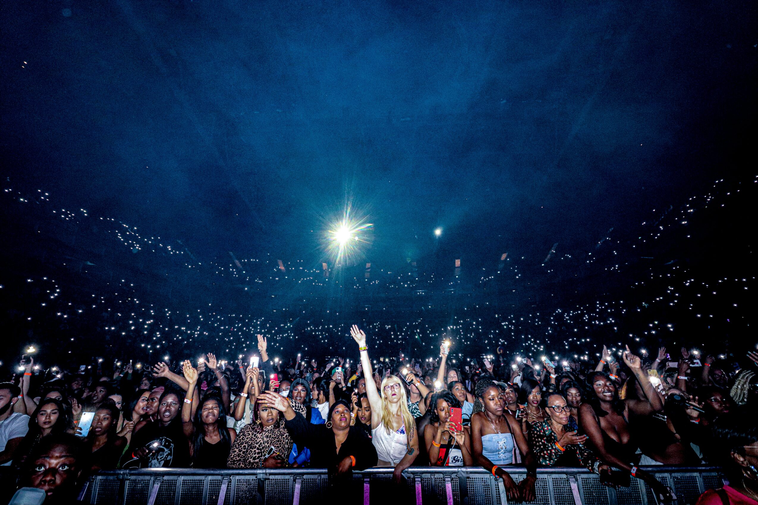 Crowd of people at concert with many holding up lights, in a dark venue with bright stage lights overhead.
