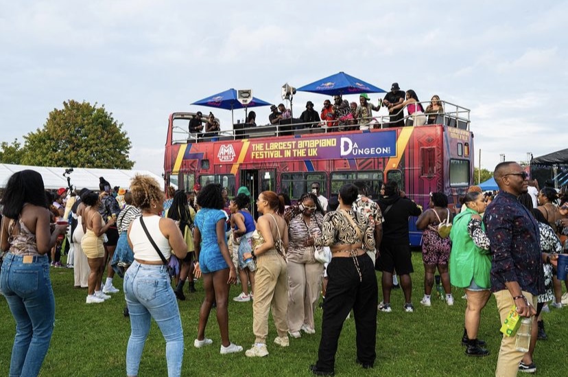 Crowd gathered around a double-decker bus with a rooftop stage and banners, outdoors on a grassy field, with trees and sky.