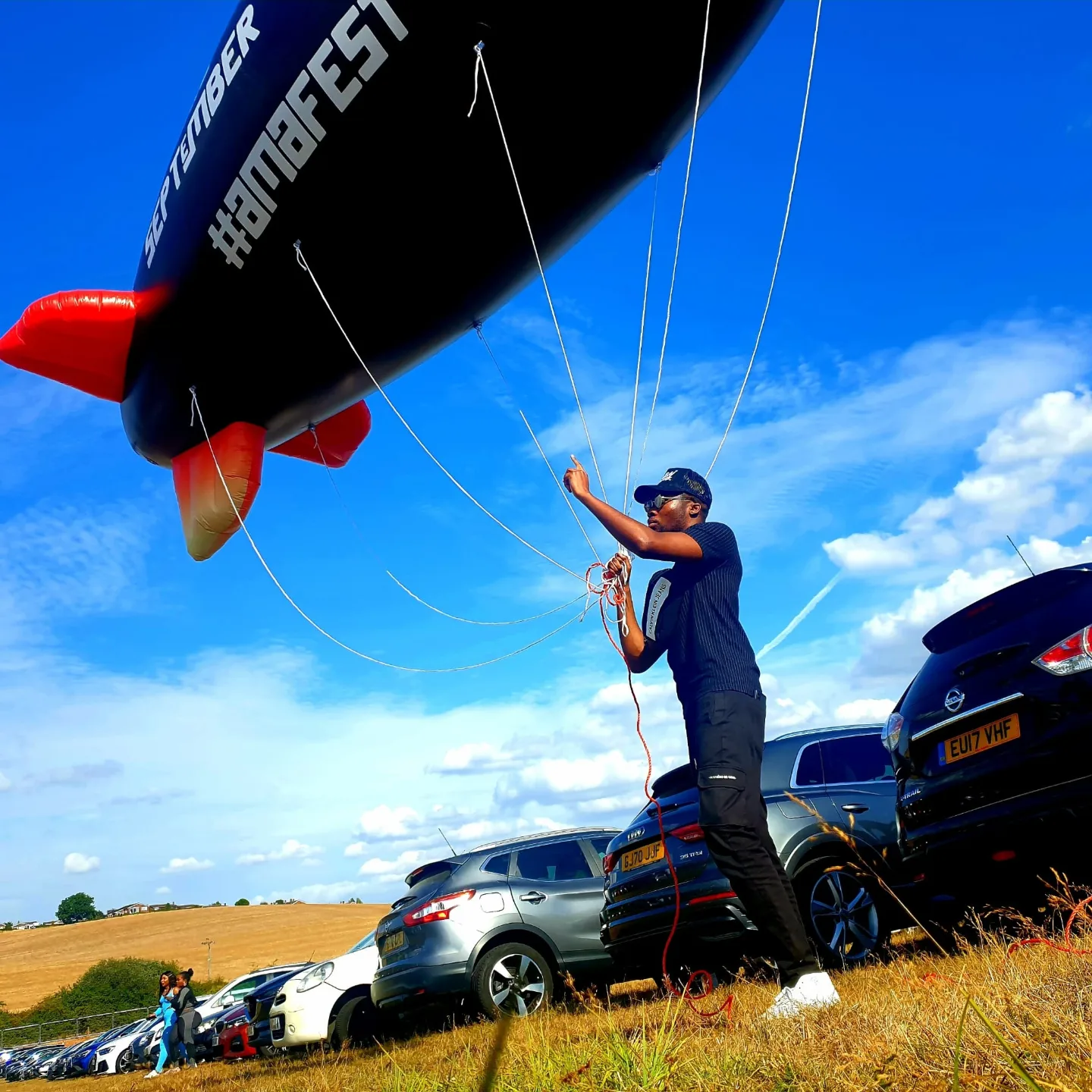 Person holding ropes near parked cars, preparing for a hot air balloon flight against a blue sky with clouds.