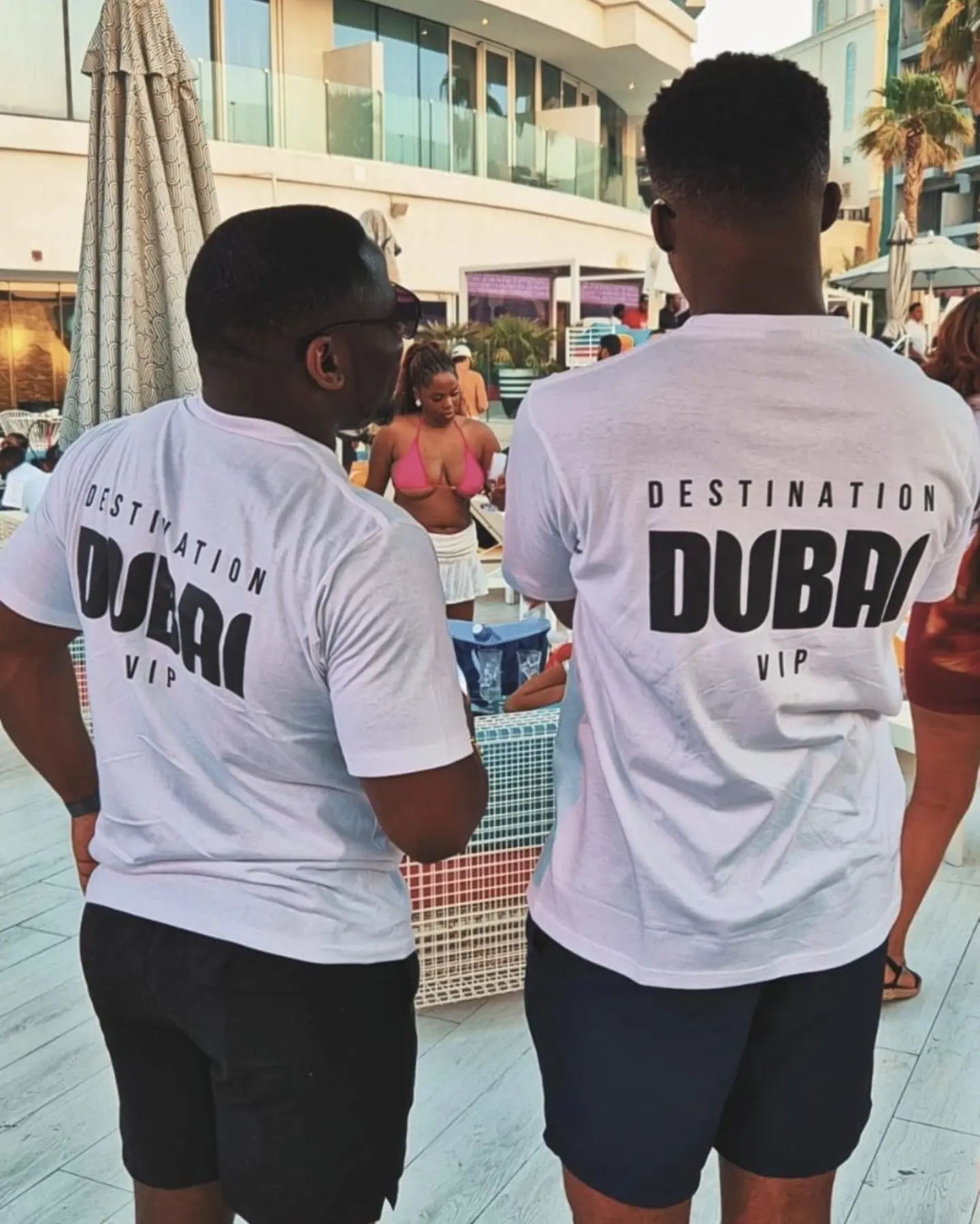 Two people wearing white T-shirts with 'Destination Dubai' printed on the back, standing outdoors near a pool or beach area.