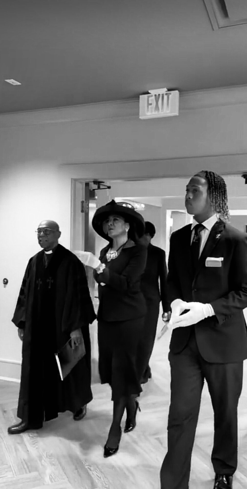 Three people walking indoors, one woman in a hat and two men in suits, with an exit sign above.