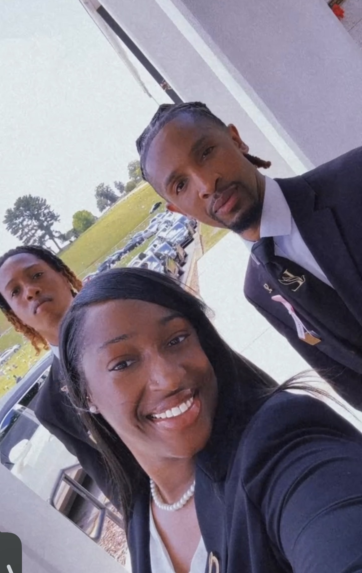 Three people smiling outdoors, two women and one man, with cars and trees in the background.
