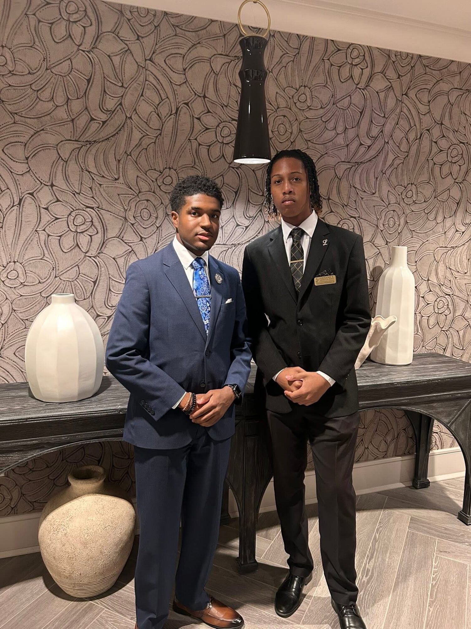 Two young men in formal suits stand indoors against a patterned wall with decorative vases and a black sculpture behind them.