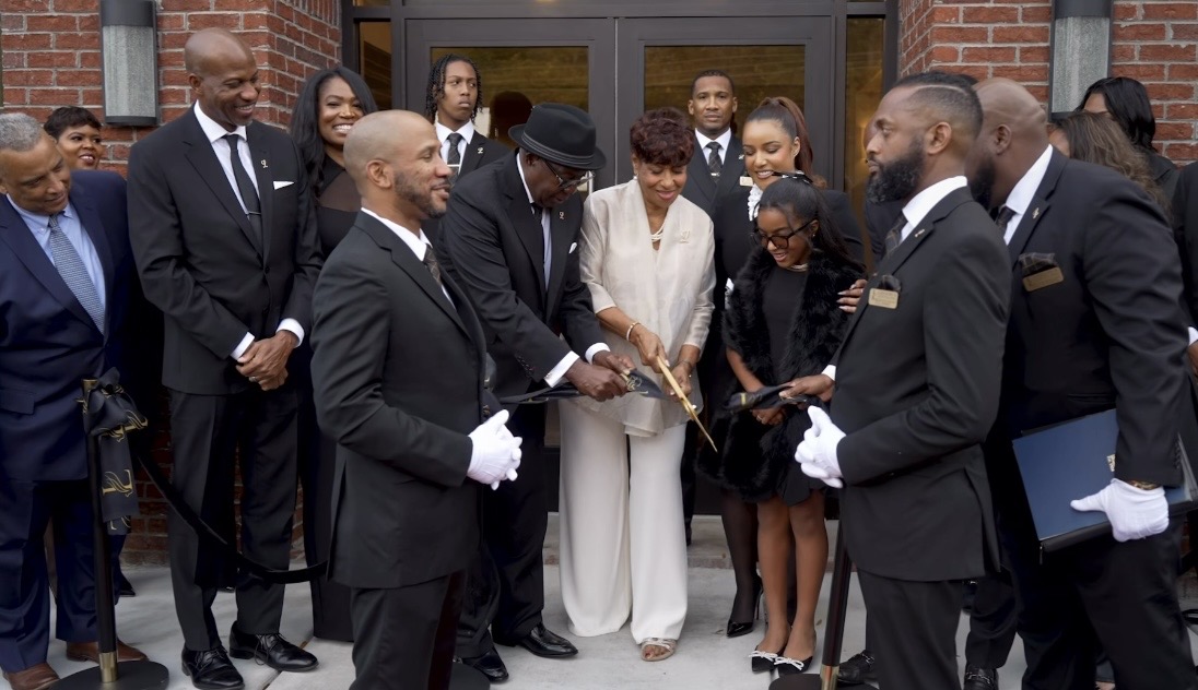 Group of people in formal attire gathered outside a building, participating in a ceremonial event, with some holding a plaque or document.