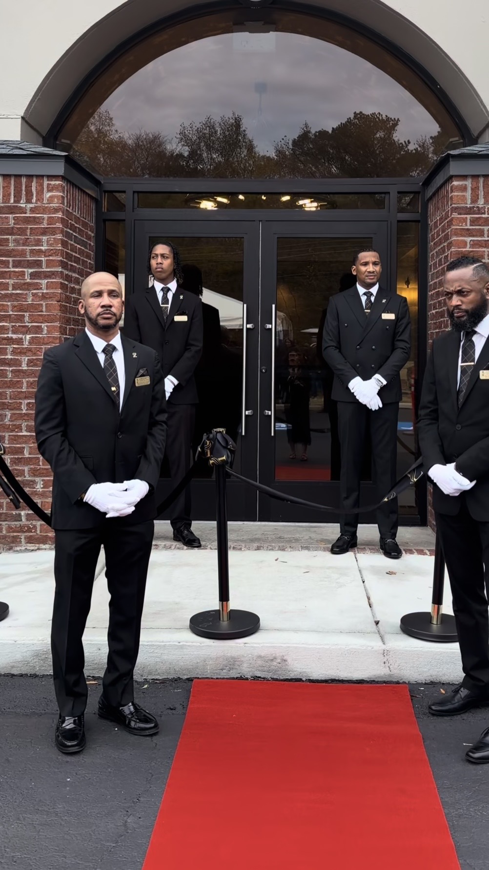 Four men in black suits and white gloves stand outside a building with a red carpet, holding ceremonial items.