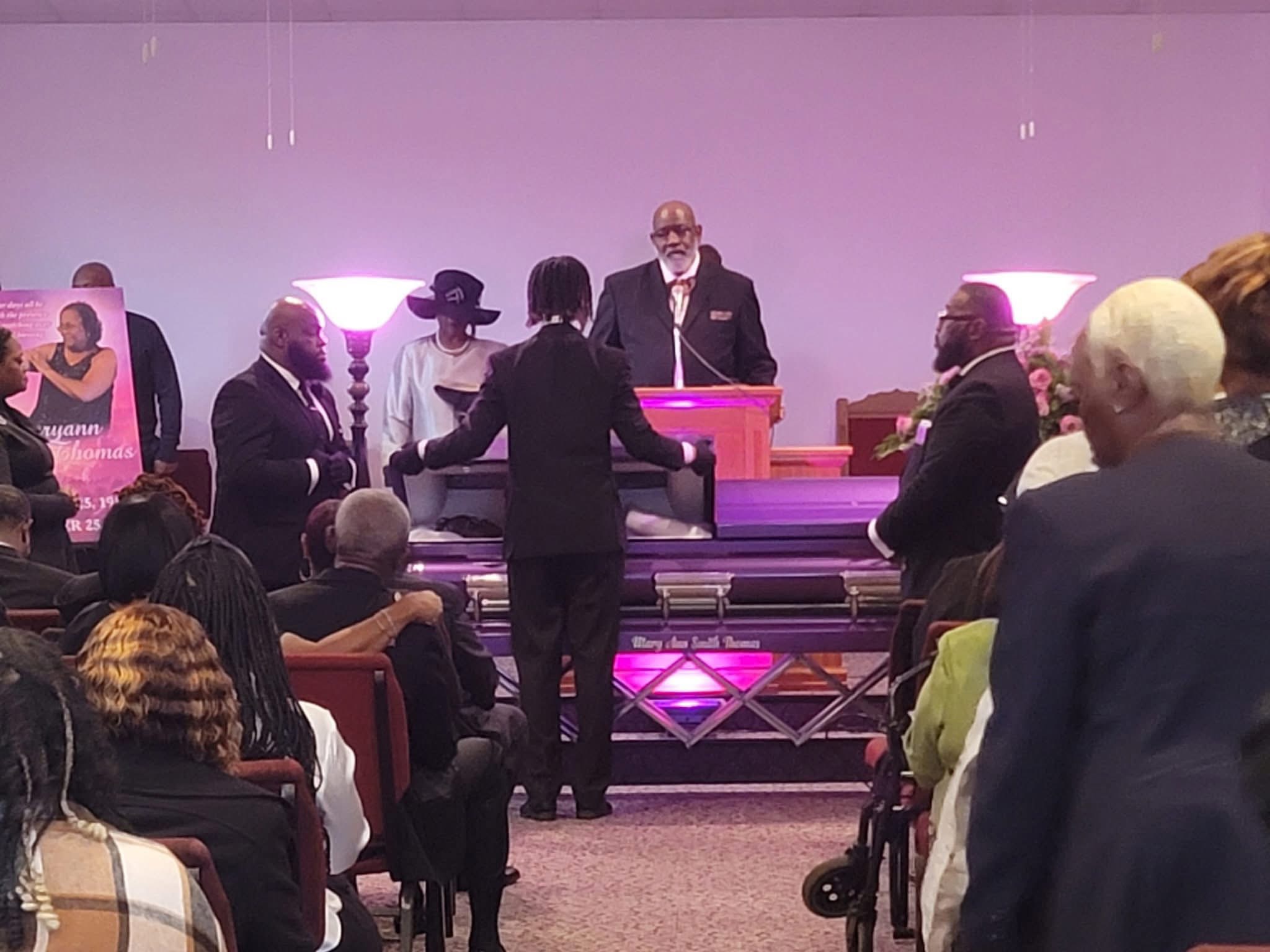 A man stands at a podium on stage with people seated and standing around, audience watching, stage lit with purple lights.