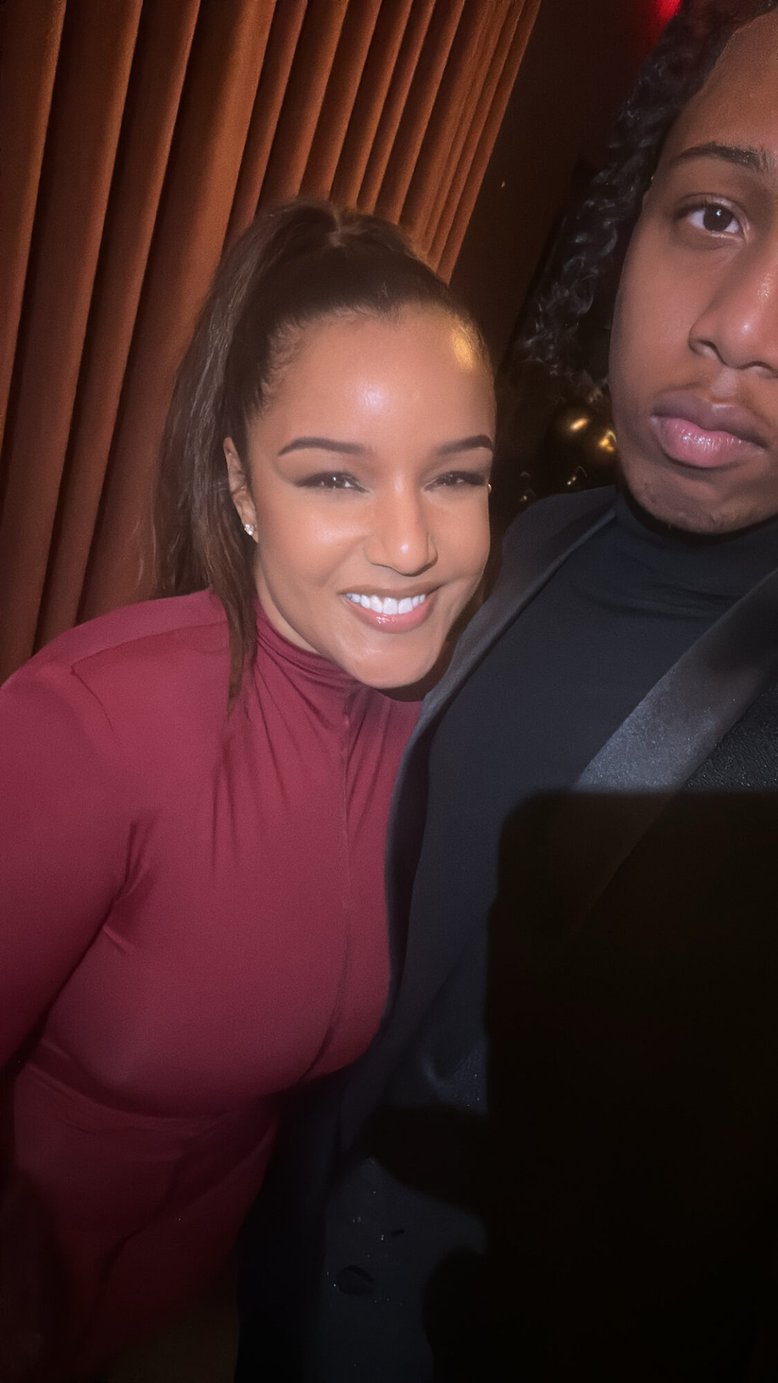 A smiling woman with long hair in a ponytail wearing a maroon top, standing next to a person with braided hair in a black shirt.