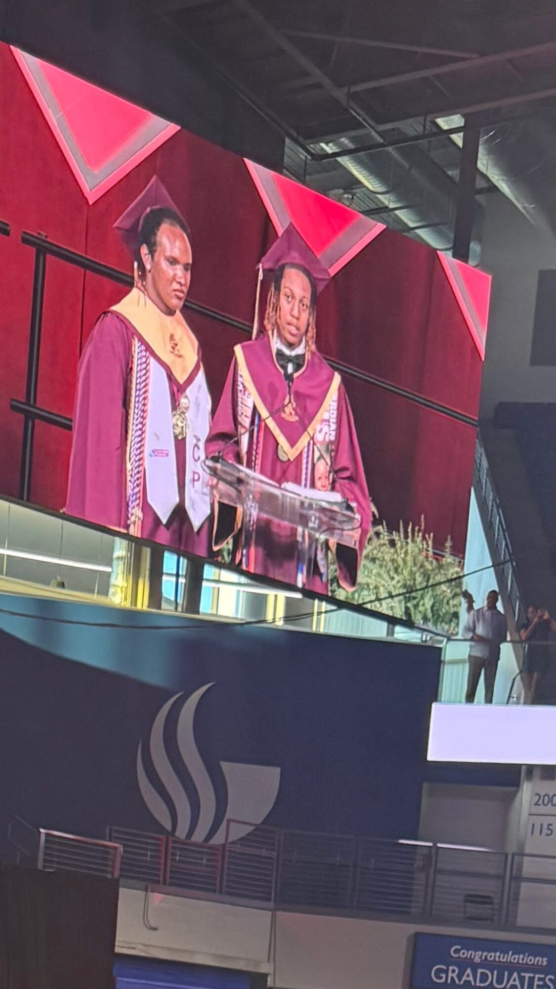 Two women in graduation gowns and caps stand on stage with a red background and a large screen behind them.