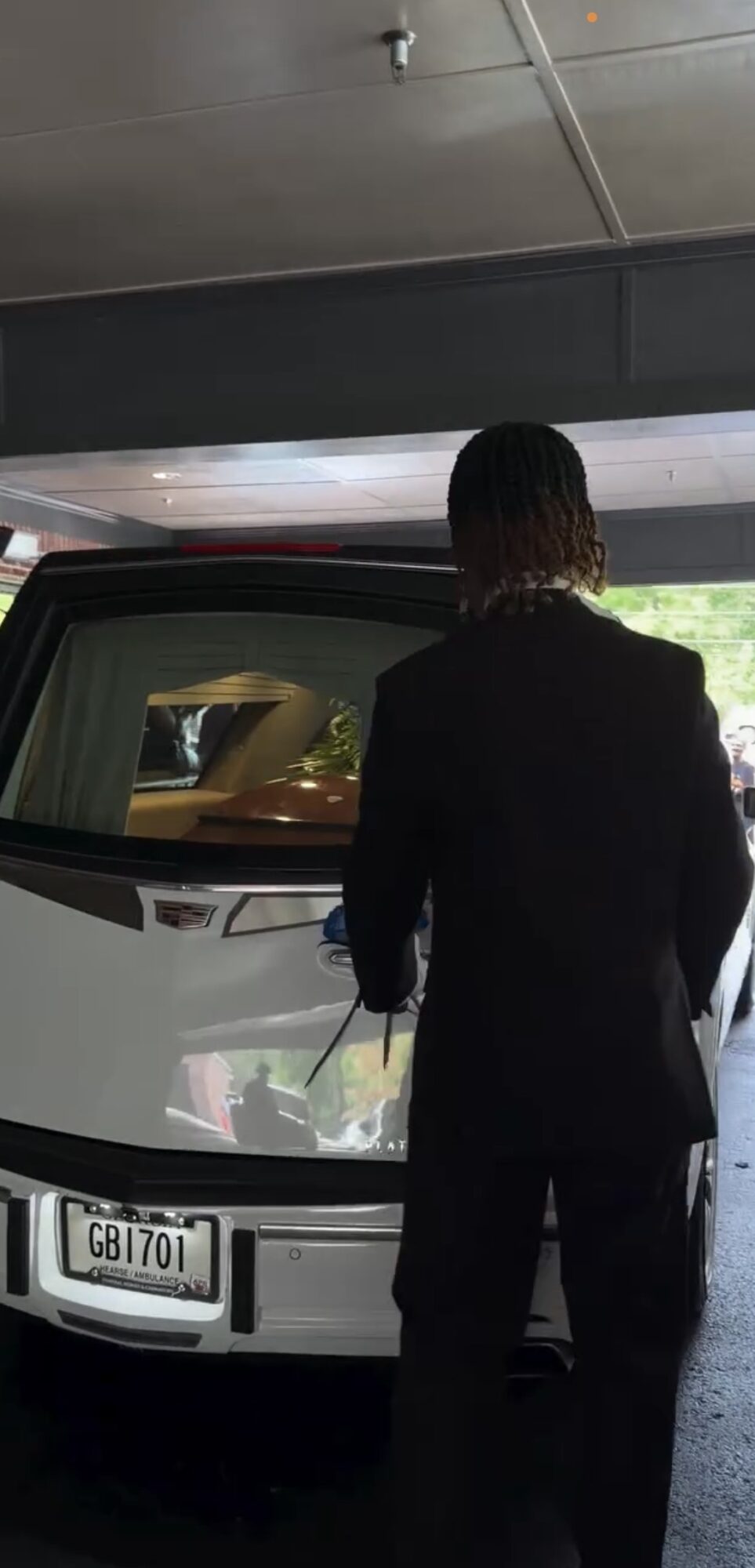 Person with dark hair in a suit stands in front of a white vehicle under a bridge or parking structure.