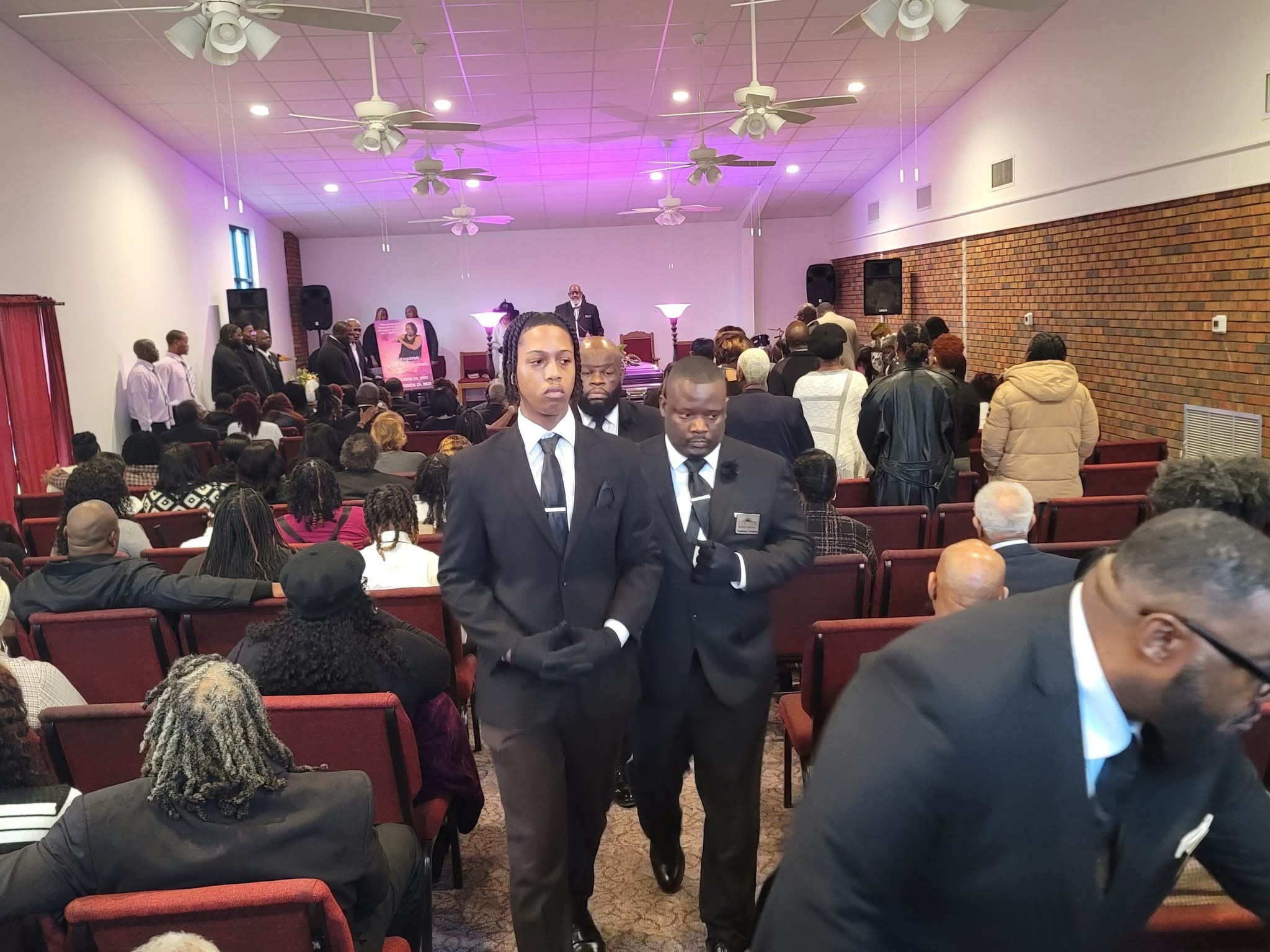 People gathered in a church or hall, some seated and some standing, with a stage and band in the background.