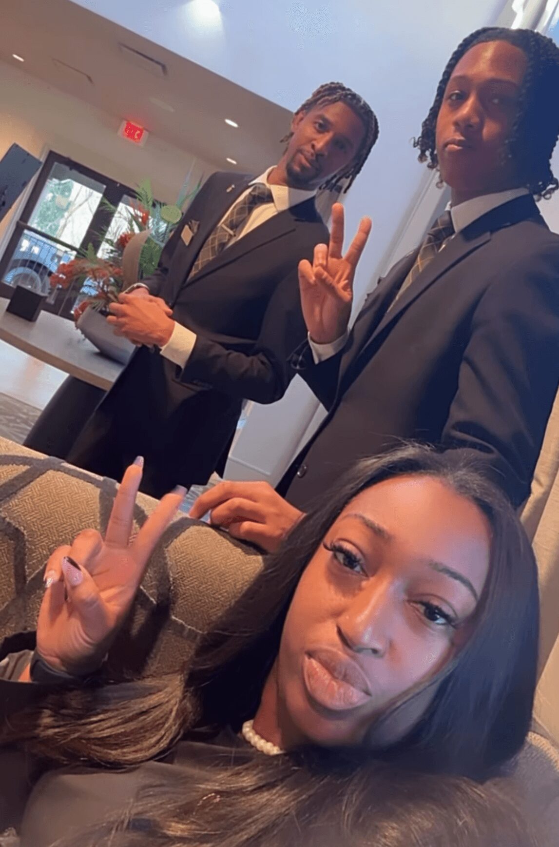 Three young people in formal attire posing indoors, two standing and one sitting, making peace signs and a pout.