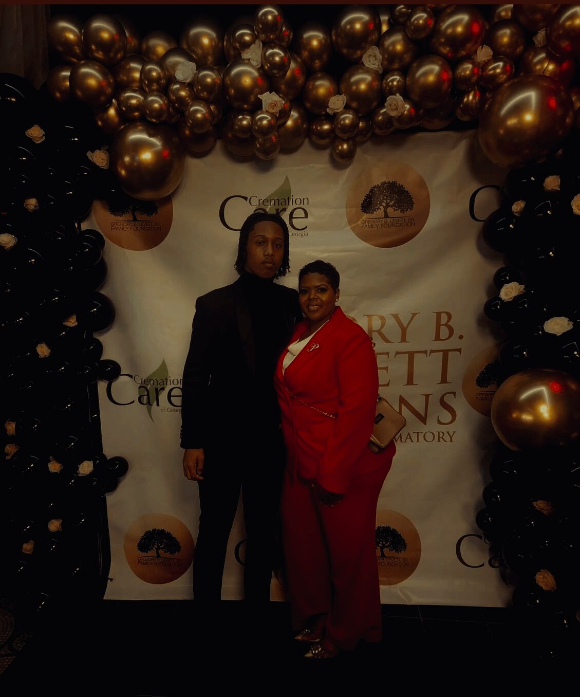 Two women standing in front of a decorated backdrop with gold and black balloons, smiling at the camera.