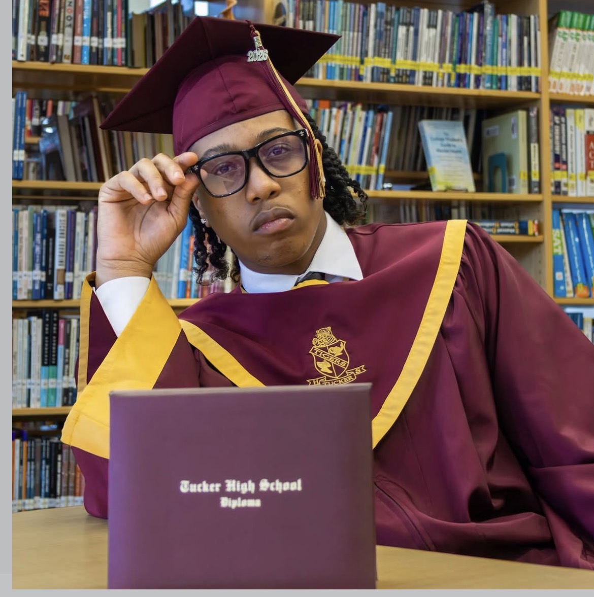 Person in graduation gown and cap sitting at a table in a library, looking at a diploma. Bookshelves in background.