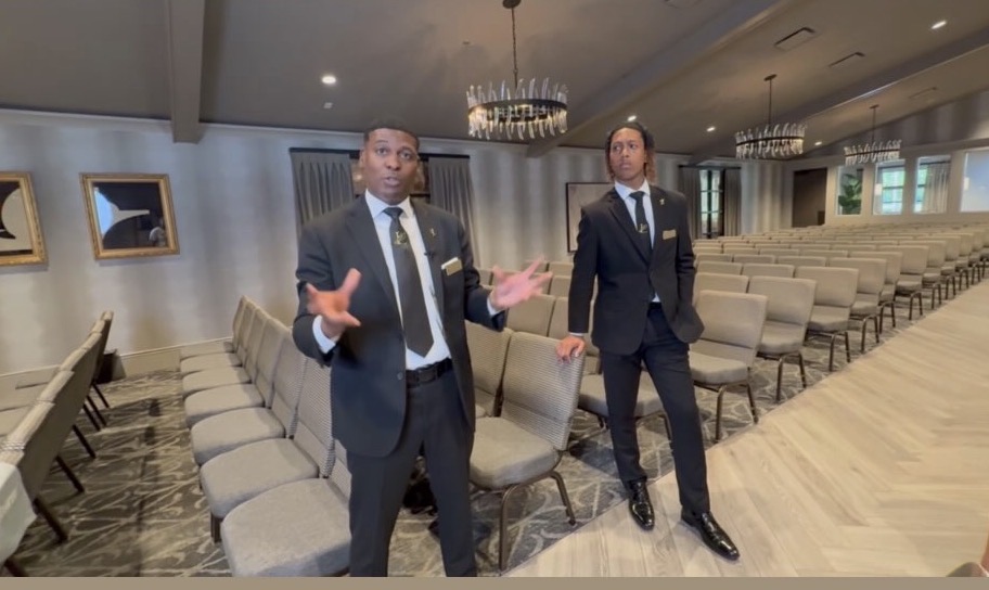 Two men in suits standing in an empty conference room with rows of chairs, chandeliers, and large windows.