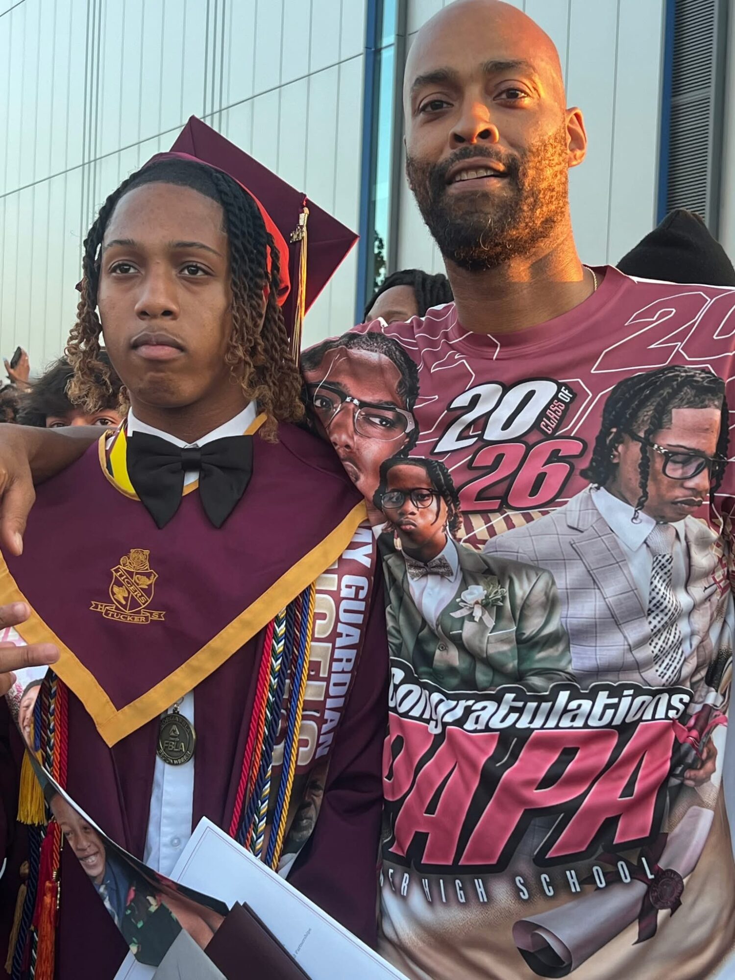 A young person in graduation attire with a maroon gown and cap, standing next to an adult man, both outdoors.