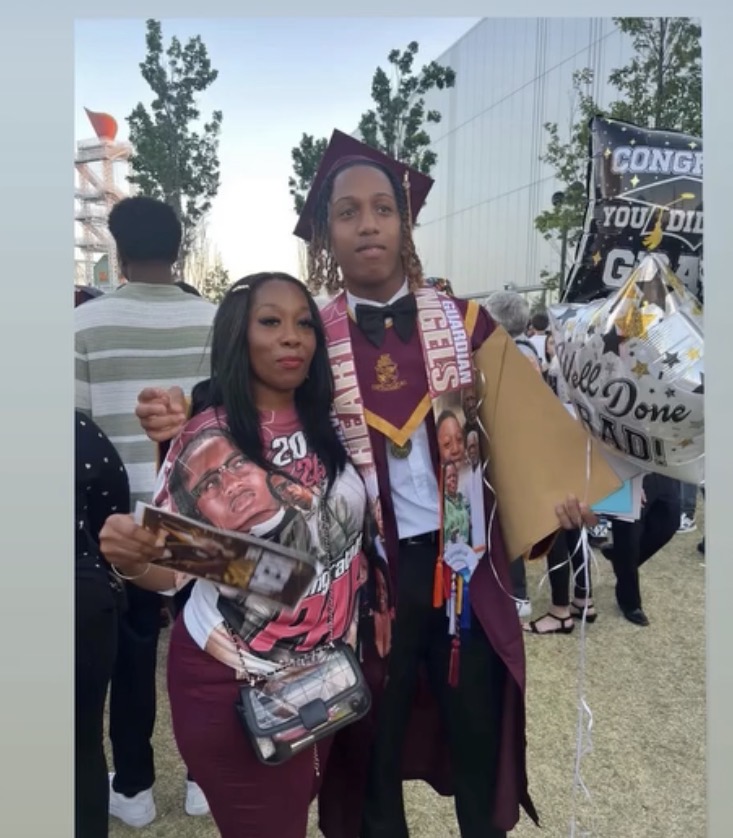 Two people at graduation, one in cap and gown holding balloons, the other holding a magazine, outdoors with trees and people in background.